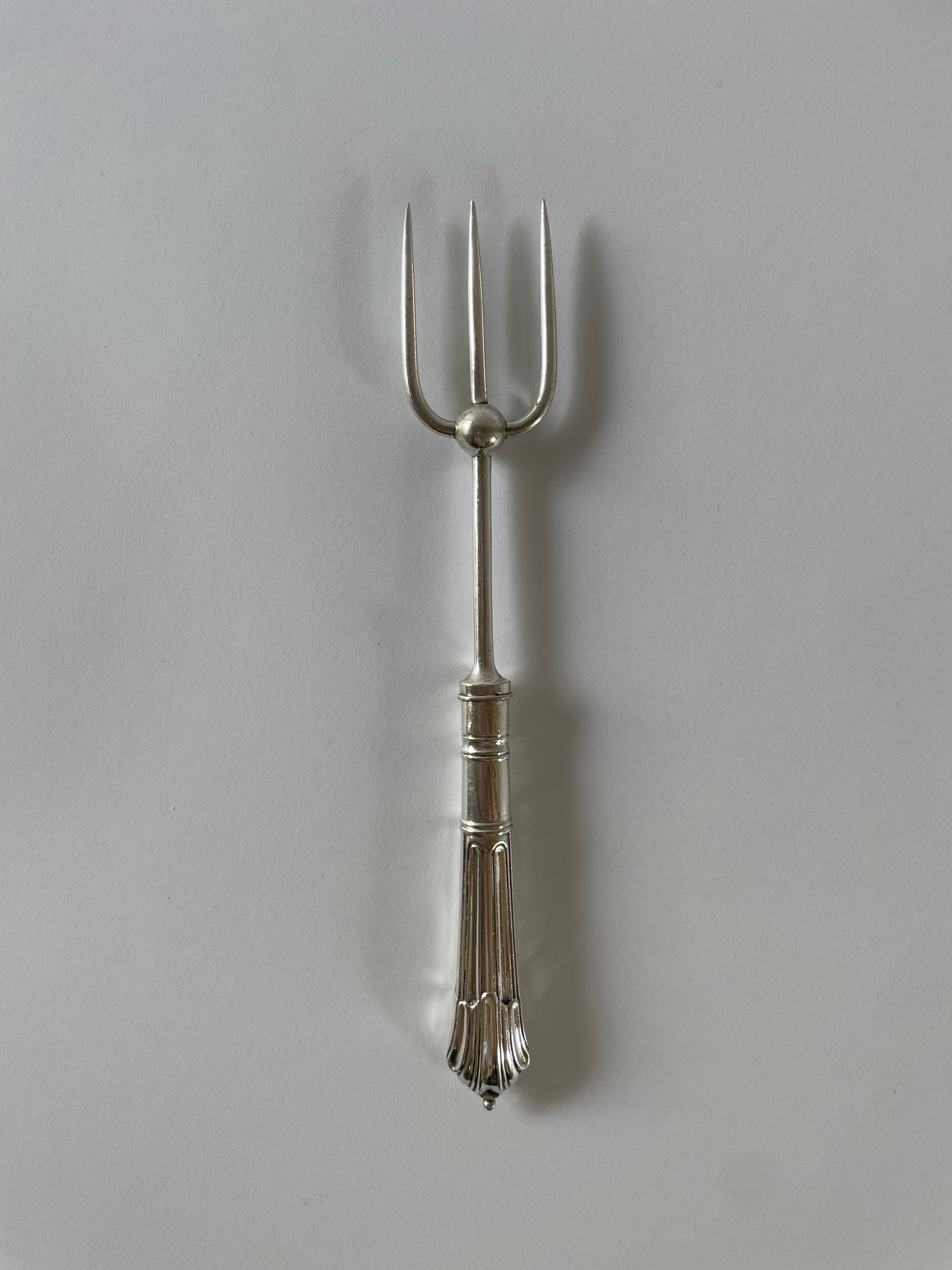 The Maud Vaughan Antique Silver Plated Bread Fork, designed in a trident style with an ornate handle adorned with intricate decorative engravings reminiscent of Victorian etiquette, is set against a plain white background. This silver-plated bread fork effortlessly elevates the presentation of fruit meats, breads, and cheeses.