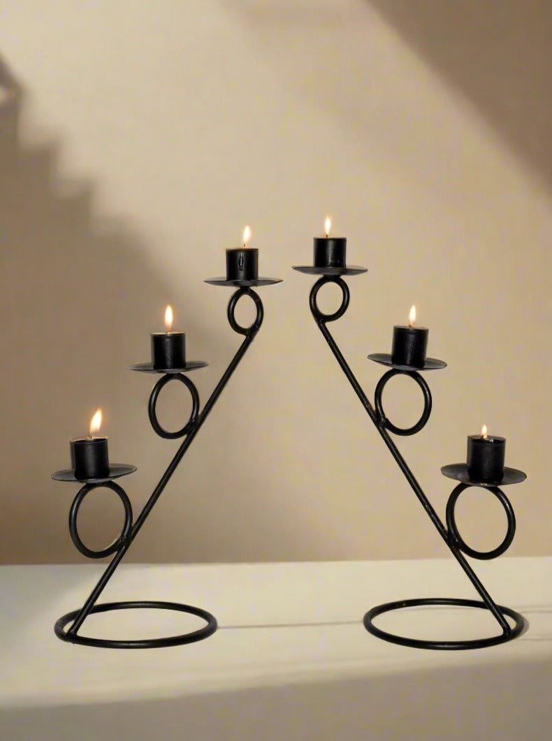 The Modernist Metal Candle Holders Pair by Break The Mould Magasin feature geometric spiral designs, each cradling three burning black candles. Symmetrically arranged on a white surface against a plain beige background, these black metal candle holders cast soft shadows on the wall, infusing the space with a touch of modernist decor.