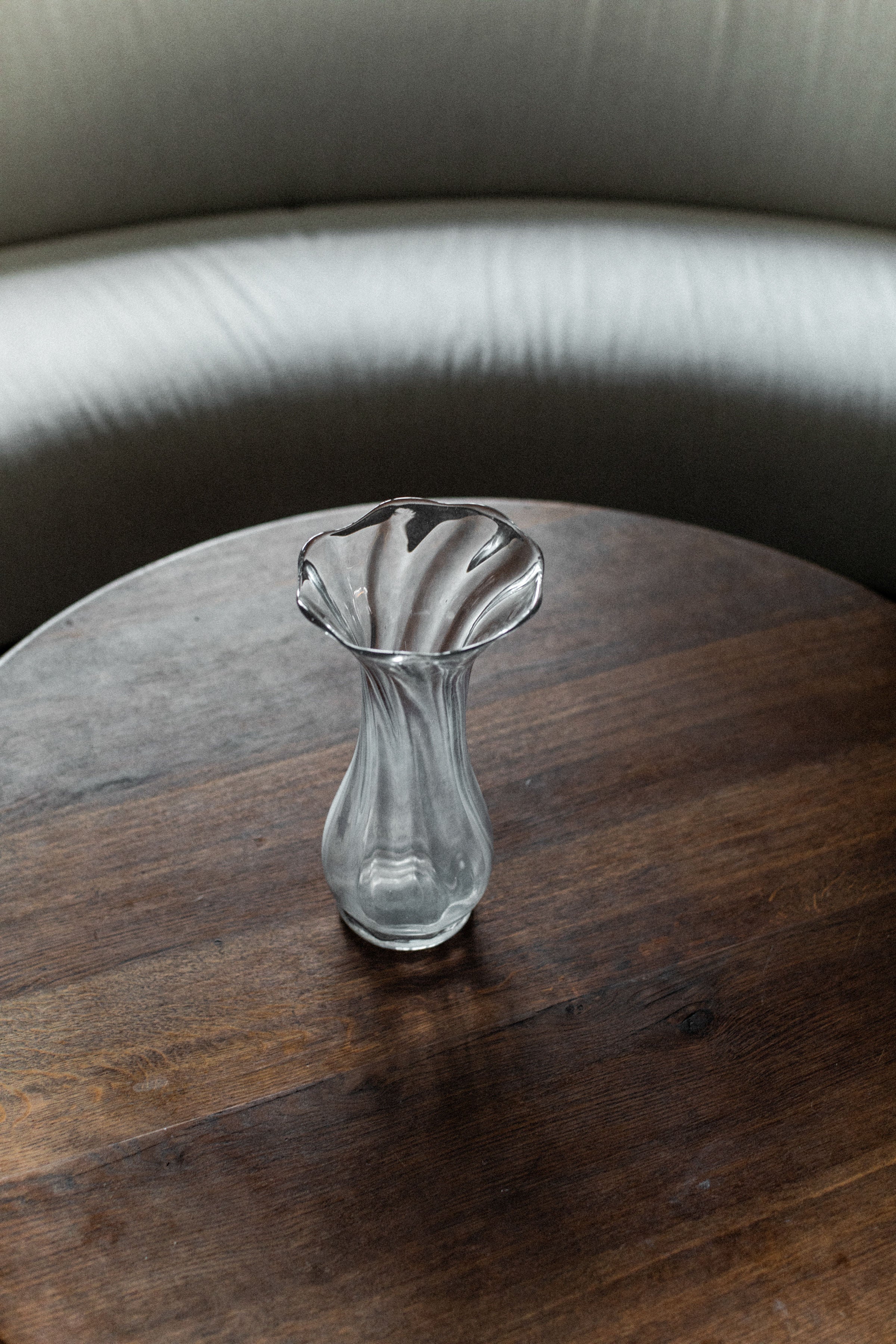 The Out For Lunch Glass Vase, featuring a wavy, flared top, is the centerpiece on a round dark wooden table. Its contemporary design pairs beautifully with the backdrop's curved light gray upholstered bench.
