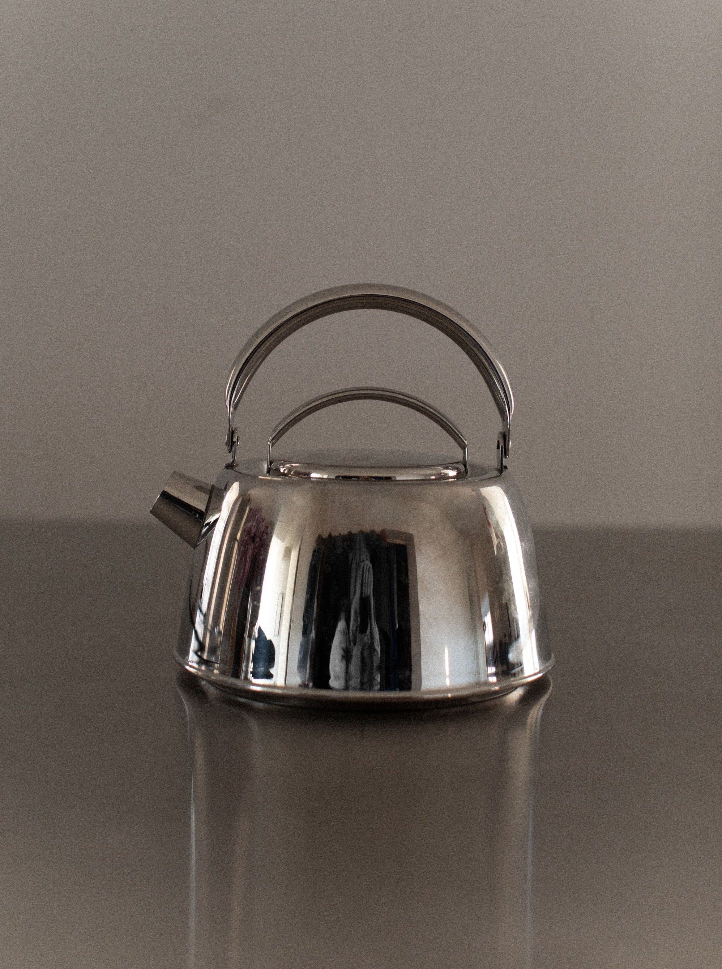 An Out For Lunch Chrome Teapot with a handle and spout sits on a reflective surface. The teapot has a polished, shiny finish that reflects parts of its surroundings, adding to its aesthetic appeal. The background is neutral and uncluttered, enhancing the refined dining experience.