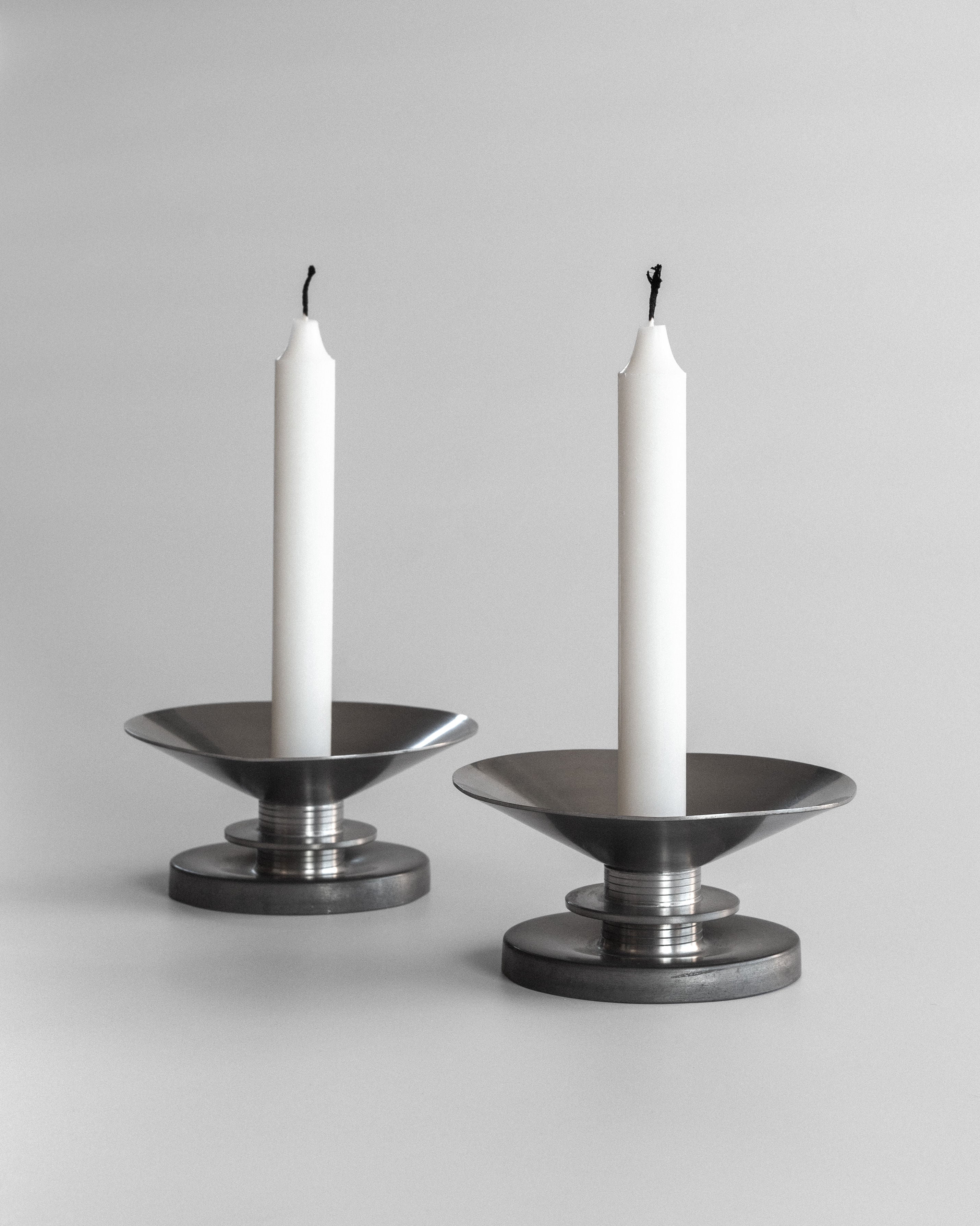 Two unlit white candles are placed in Spigel's Modernist Candleholders by Sylvia Stave 1930s on a gray background. Resembling pewter candlesticks, these stainless steel holders have a sleek, minimalist design with a wide dish and round base, echoing 20th-century decorative arts.