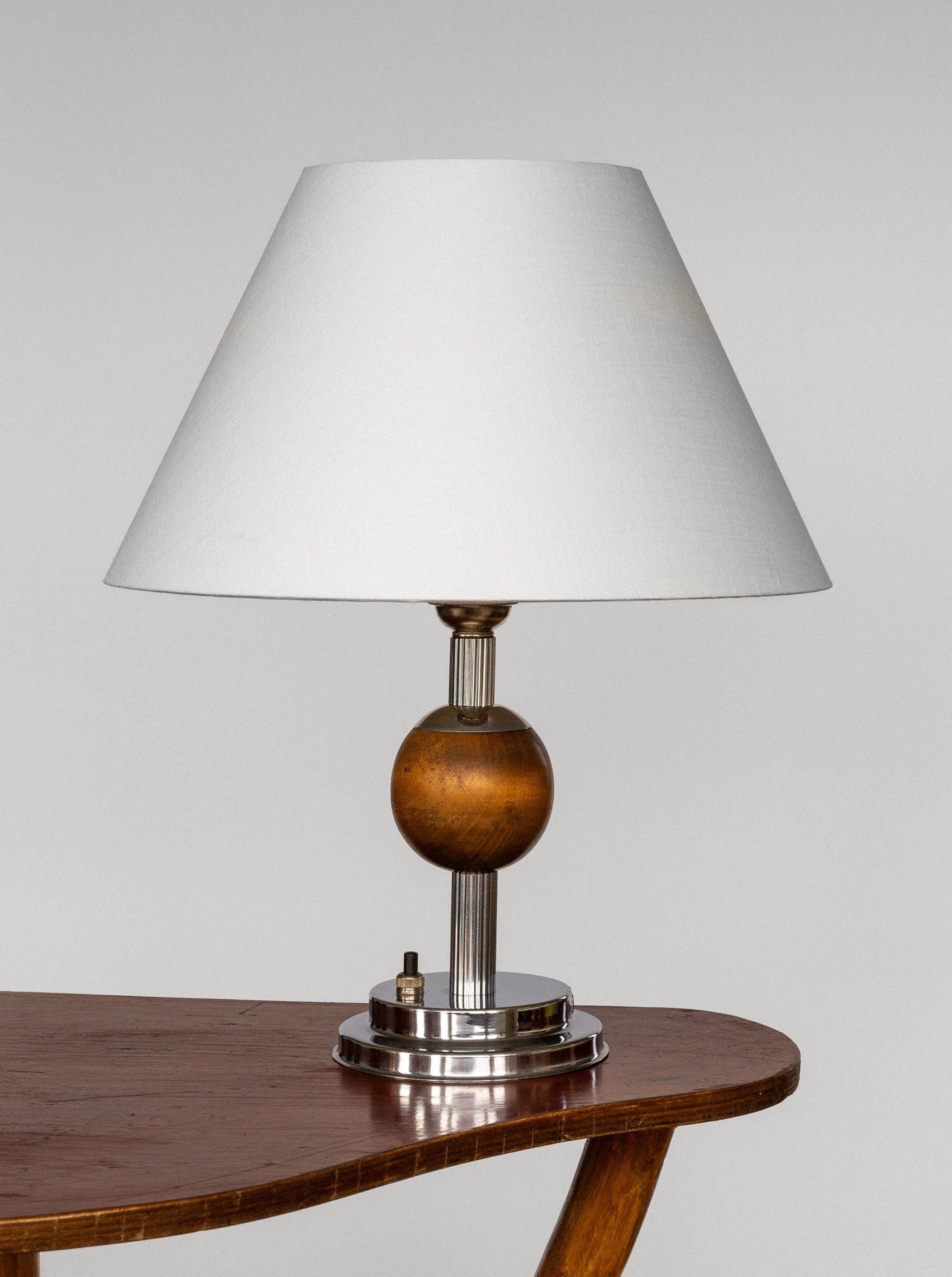 A Swedish Art Deco Metal and Wood Lamp from the 1930s by Spigel, featuring a white conical shade, wooden spherical base, and cylindrical metallic support, is placed on a wooden surface with rounded edges. The lamp's modern and minimalist design evokes the elegance reminiscent of the Art Deco era.