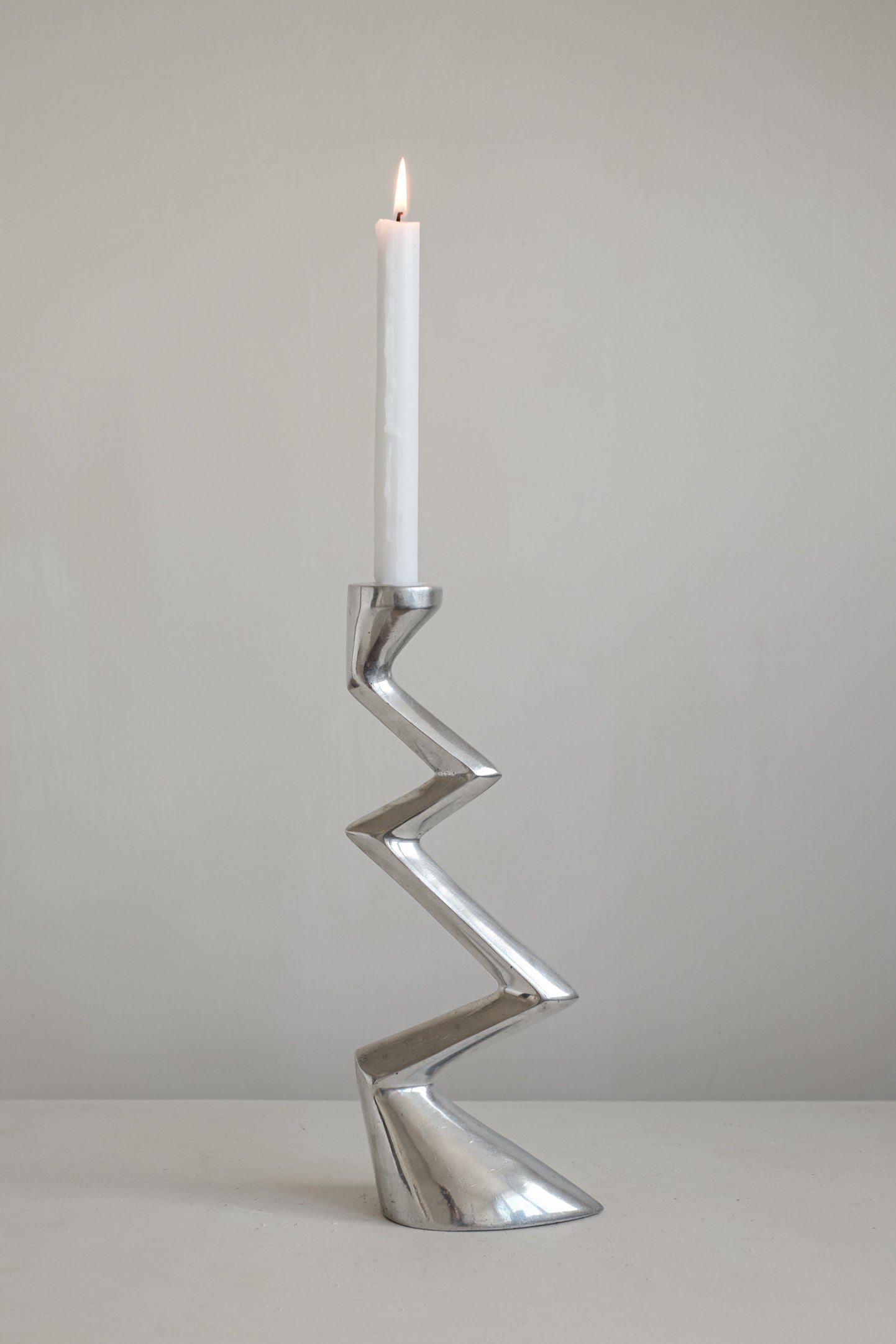 A white candle stands in the septembre studios Matthew Hilton ZigZag Candle Holder Large, featuring an angular silhouette, perfect for contemporary interiors, set against a plain light gray background.