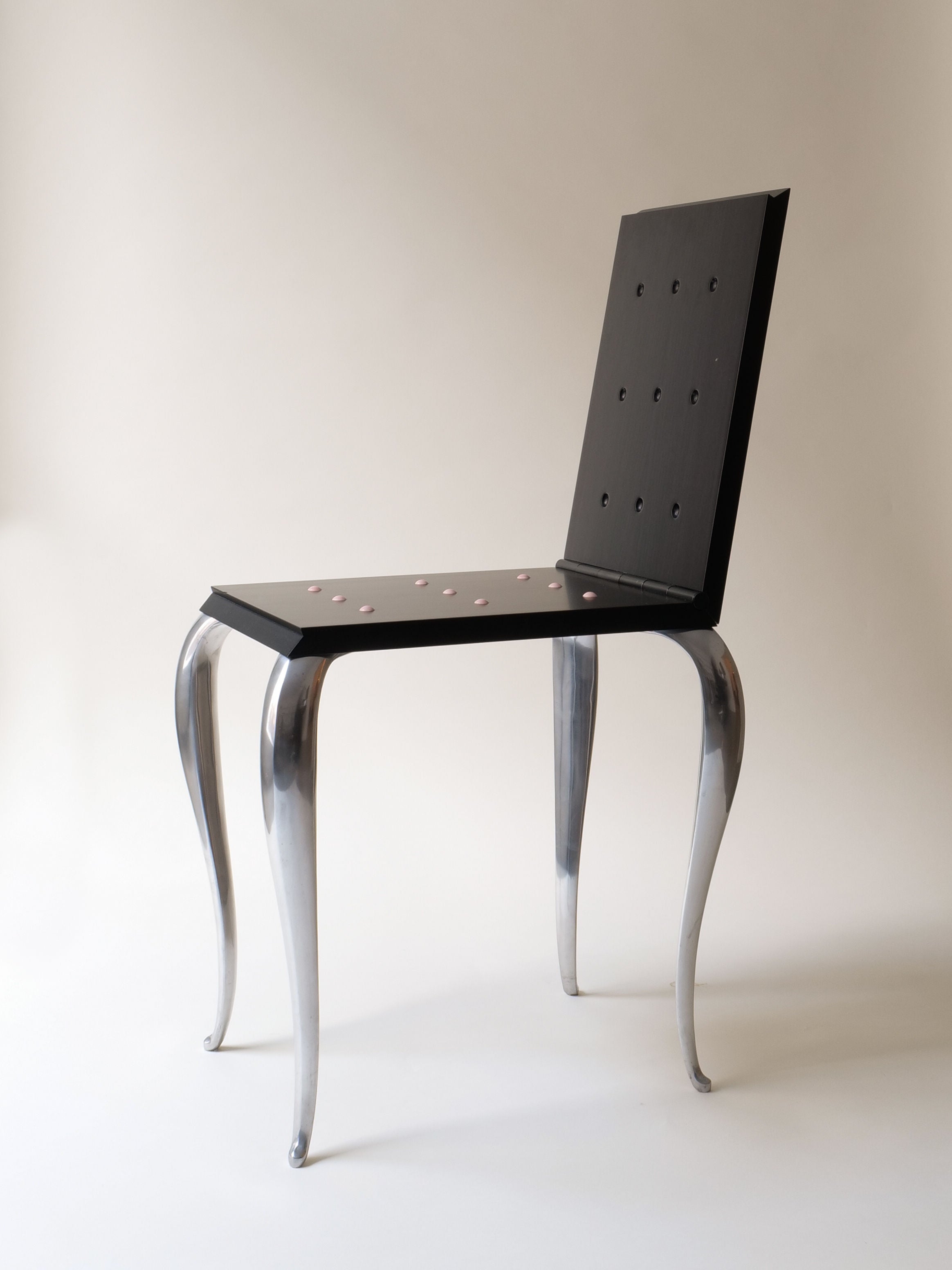 The "Lola Mundo Chair" by Philippe Starck for Collection apart, designed in 1984, features a sleek black seat and backrest with dots, reminiscent of Starck's aesthetic. It has four shiny, curved metal legs set against a plain, light-colored backdrop.