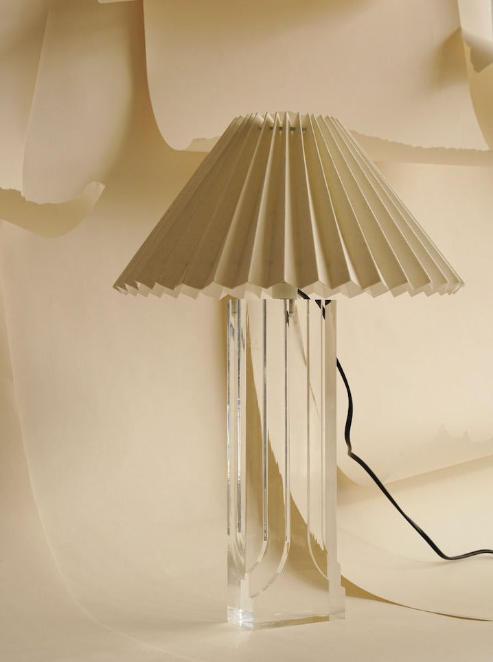 An Art Deco "Ghost" Lamp by Les Objets Amsterdam features a transparent base and a pleated beige lampshade set against a backdrop of cream-colored, textured paper. A black electrical cord is partially visible, extending from the base of this classic piece.