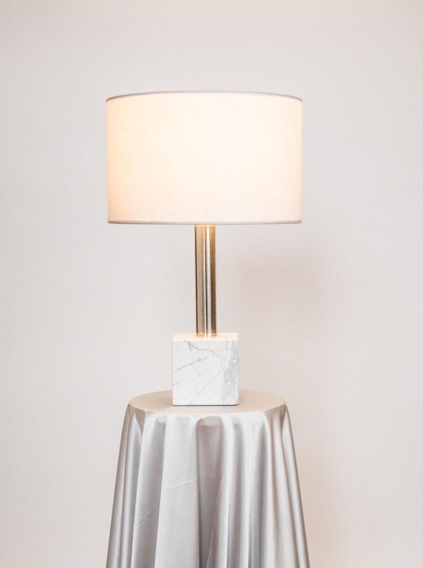 A stylish Brajak Vitberg Athens Table Lamp 2 with a Carrara marble base and a sleek polished stainless steel stand, topped with a cylindrical light shade, placed on a draped table with a soft, gray fabric against