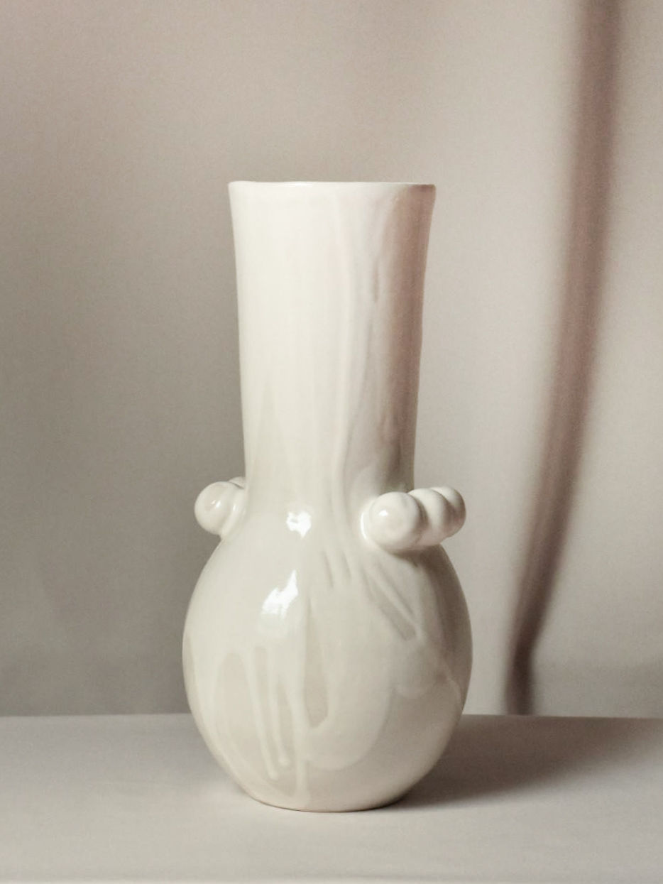 The Vase Solstice I by Landy Rakoto Ceramics is a handmade white ceramic piece with a smooth texture, bulbous base, and two side protrusions. It stands out against a light background, showcasing its minimalist elegance.