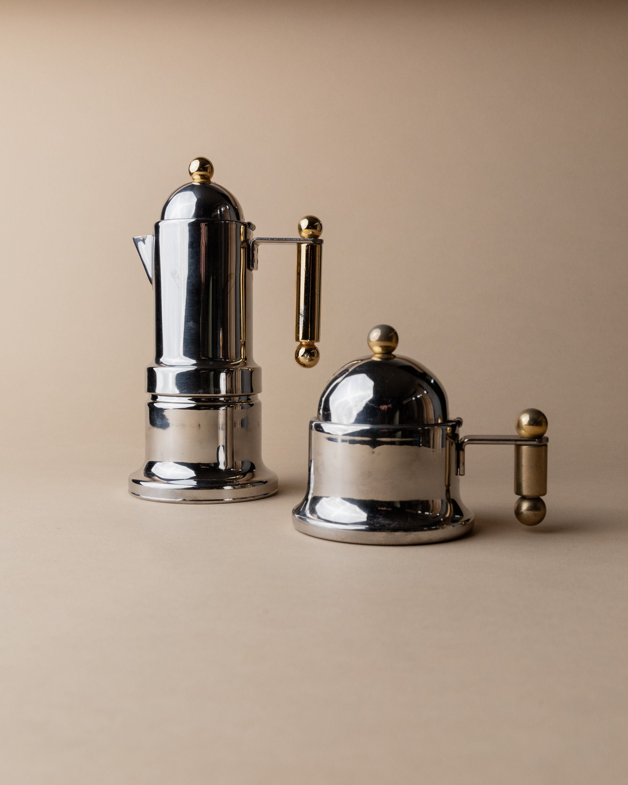 Two Coffee Set by VEV Italy 80s pieces by Bottega Jacobs feature silver and gold metal with rounded tops and handles, evoking vintage Italian mocha pots, set on a neutral beige surface against a matching backdrop.