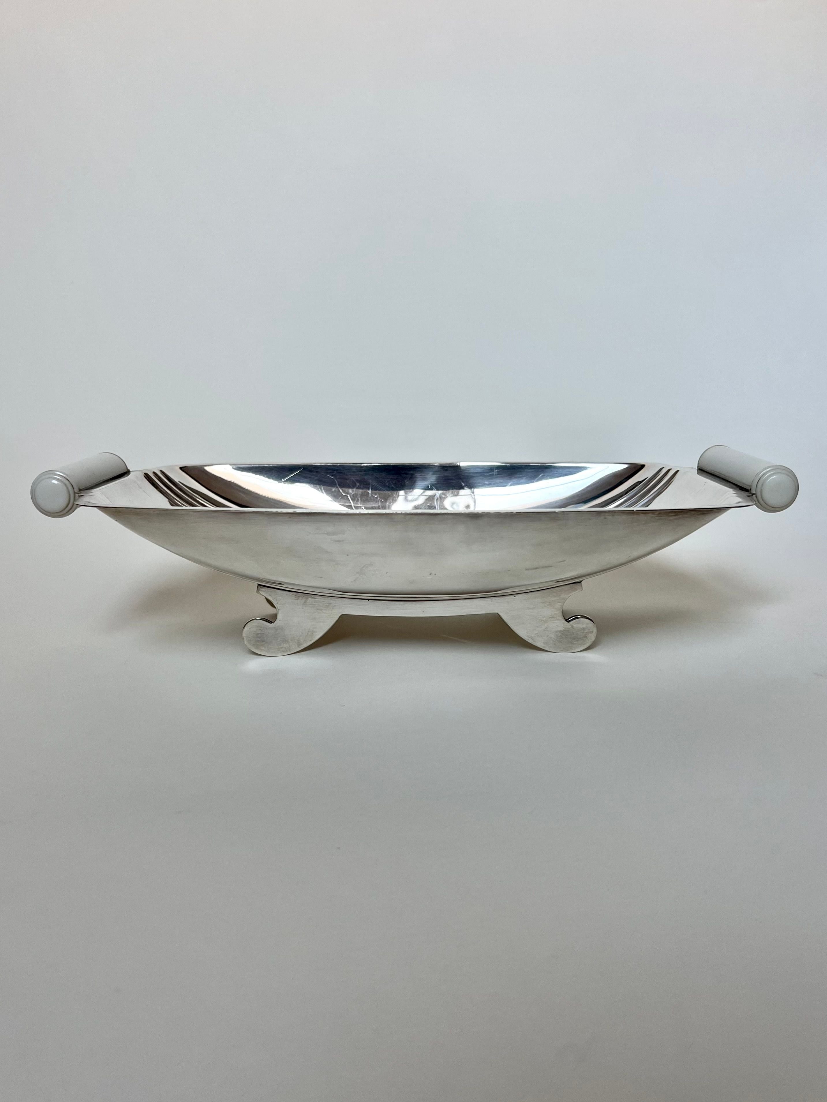 The Manushi Art Deco Bread Dish With Handles from France boasts a sleek silver design with decorative side handles and two curved feet, offering a polished surface that subtly reflects light against a plain background.