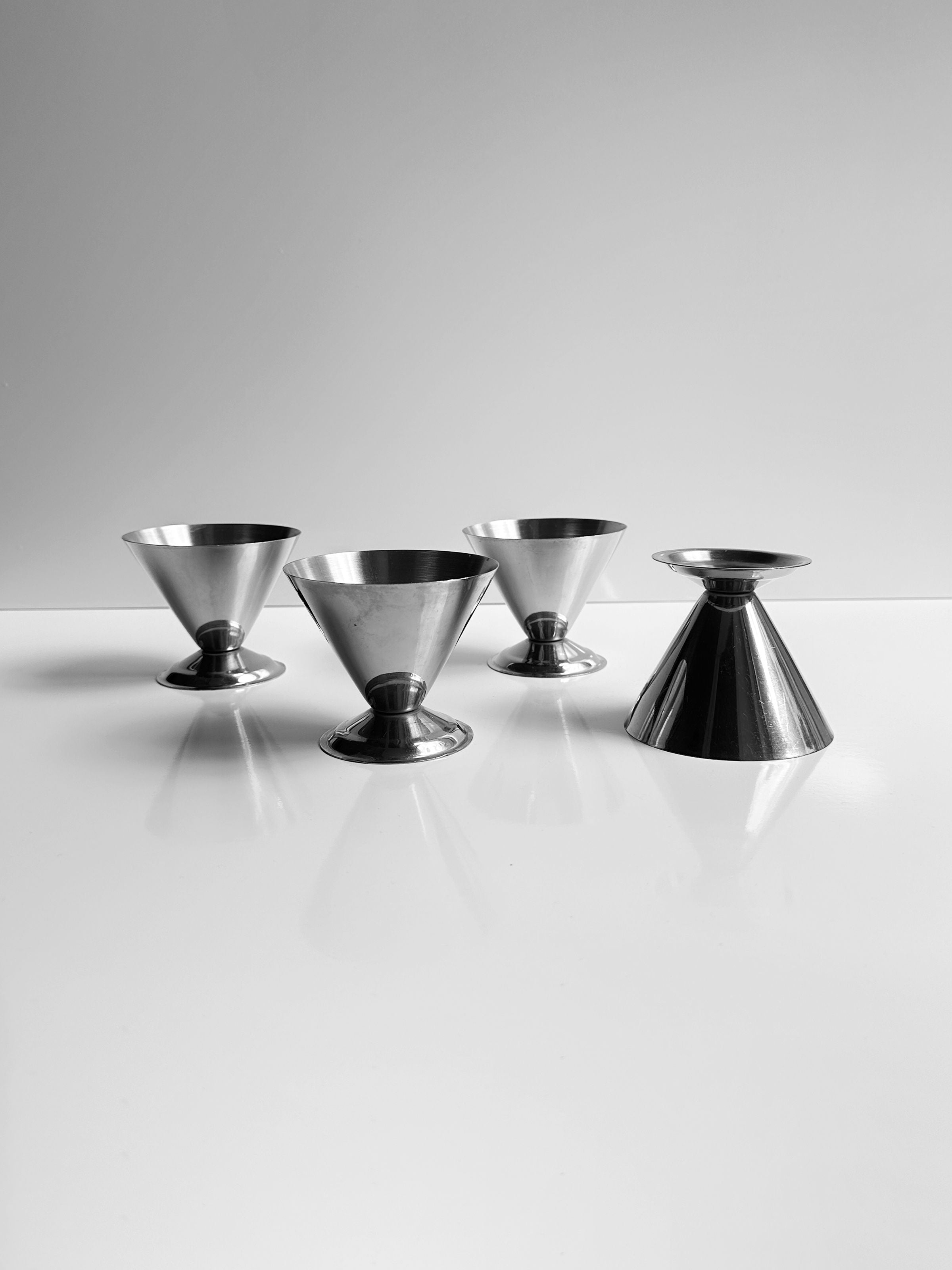 Four items from the Set of 4 Italian Stainless Steel Dessert Cups by Dodo Vintage are arranged on a glossy white surface, contrasting against a gray backdrop. Each cup, with its wide opening and diverse size, stands upright elegantly, evoking the style of 1980s Italian design.