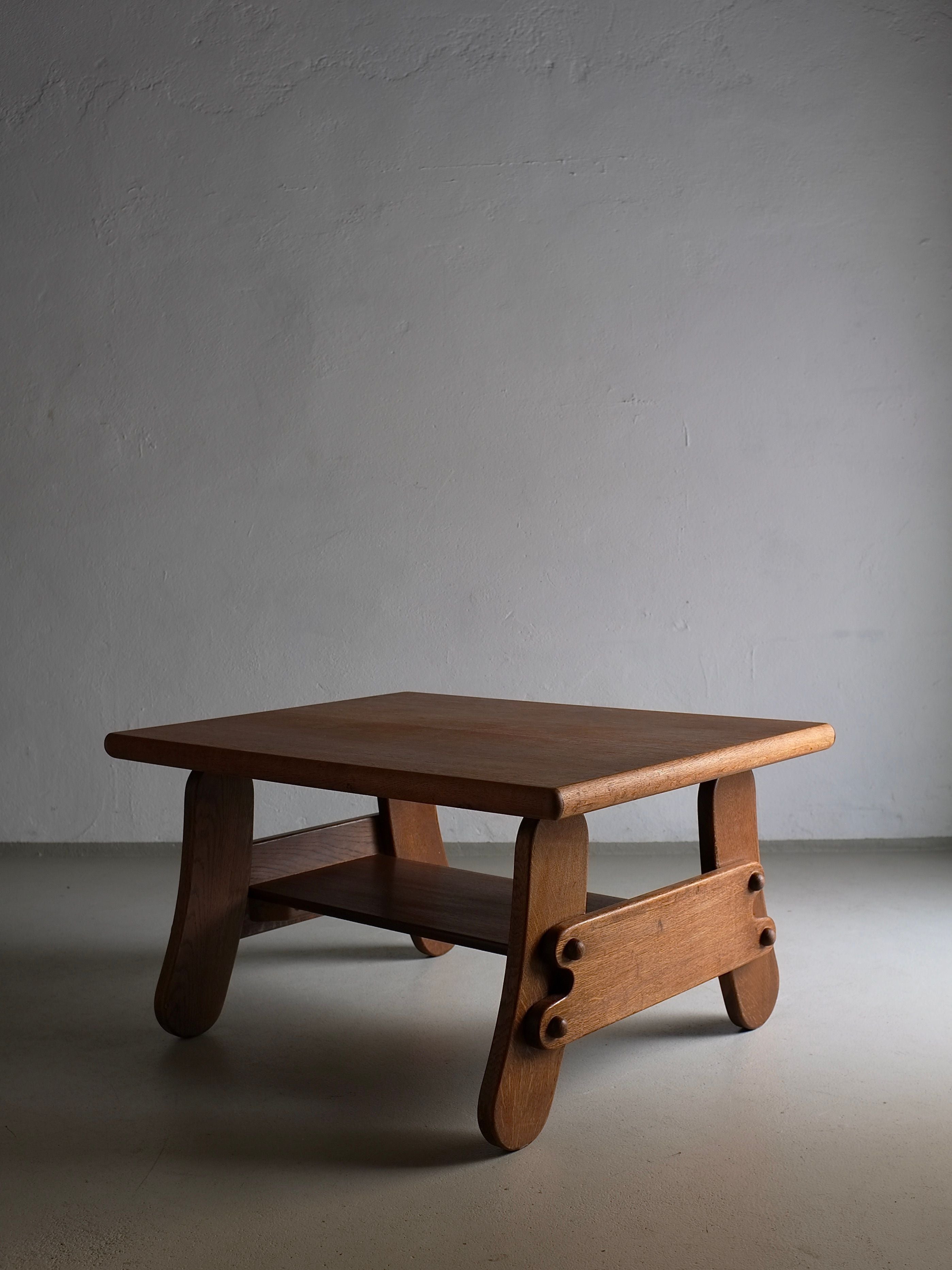 A small Brutalist Oak Coffee Table | Netherlands | 1970s with a simple, 1970s design stands against a plain, light gray wall. The table has a square top, curved legs, and a lower shelf. The lighting casts soft shadows, highlighting the table's natural wood grain and craftsmanship. This piece is from Veter Vintage.