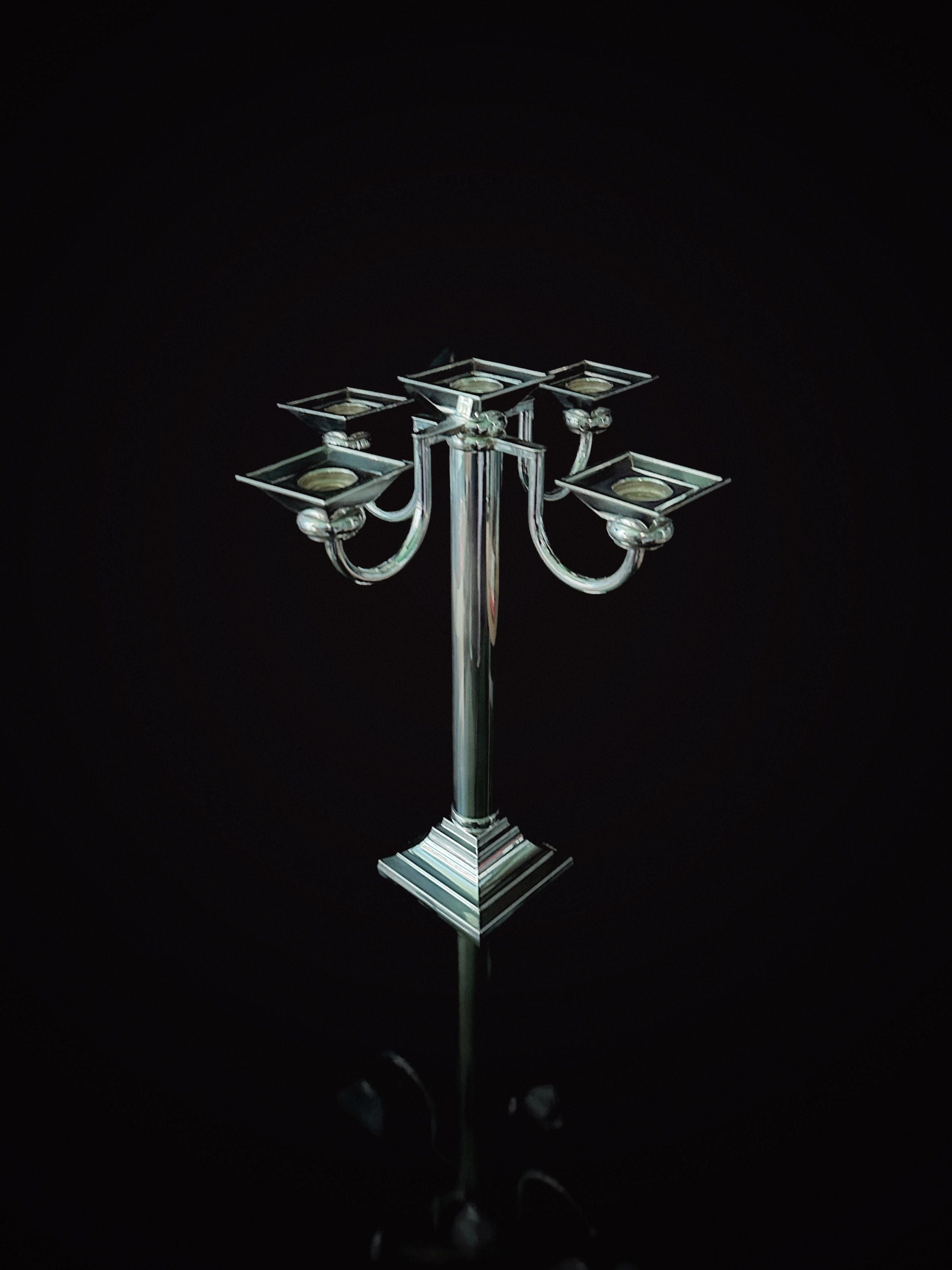 The Silver Plated Art Deco Candelholder 80s by Dodo Vintage features five intricate arms, each adorned with a square candle holder, showcasing its elegant Art Deco design. The polished surface of the candelabrum reflects light beautifully against a dark backdrop, highlighting its sophisticated and symmetrical structure akin to Fink Germany craftsmanship.