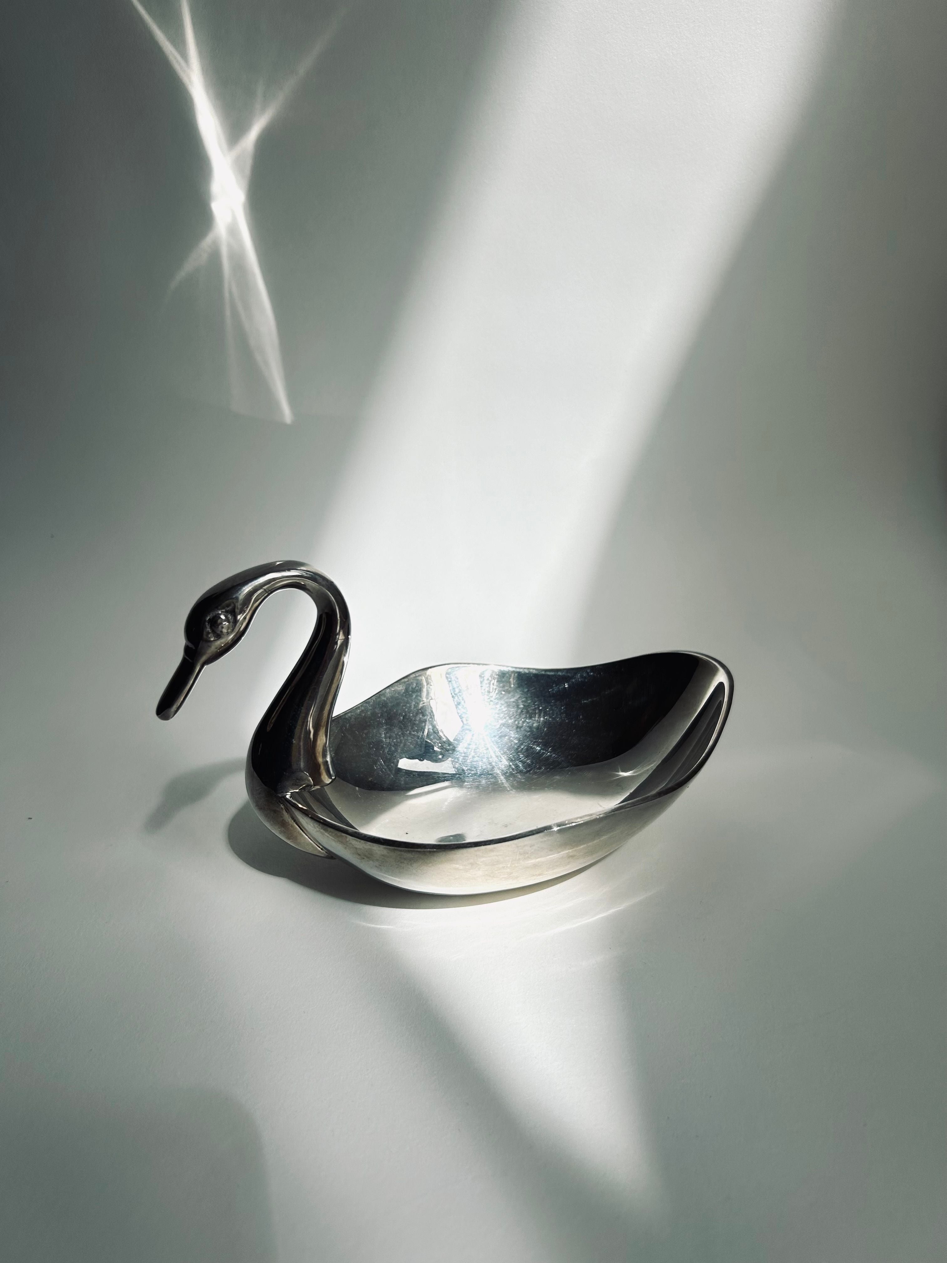 The Silver Plated Swan Dish by Flock Studio, ideal for holding candy at your party, is bathed in a ray of light against a neutral backdrop, producing a starburst effect on its glistening surface.
