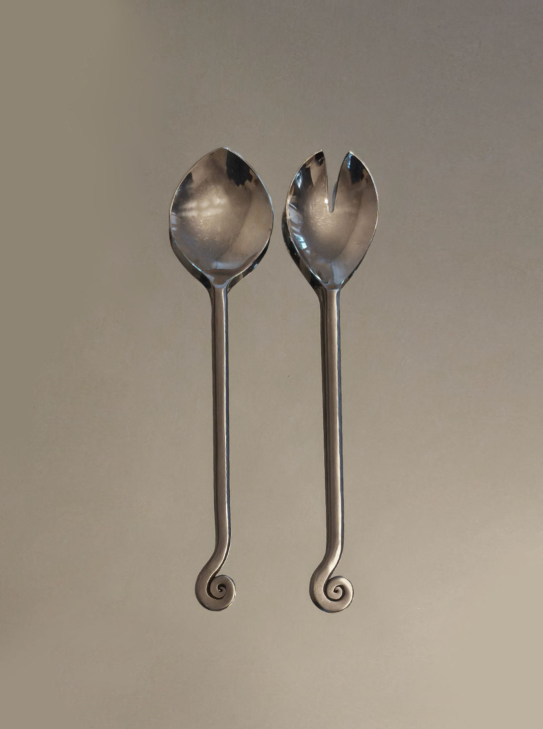 The Les Objoies Spiral Salad Servers feature long handles with spiral ends—one solid spoon, one forked—for a sculptural, decorative touch that stands out against a neutral background.