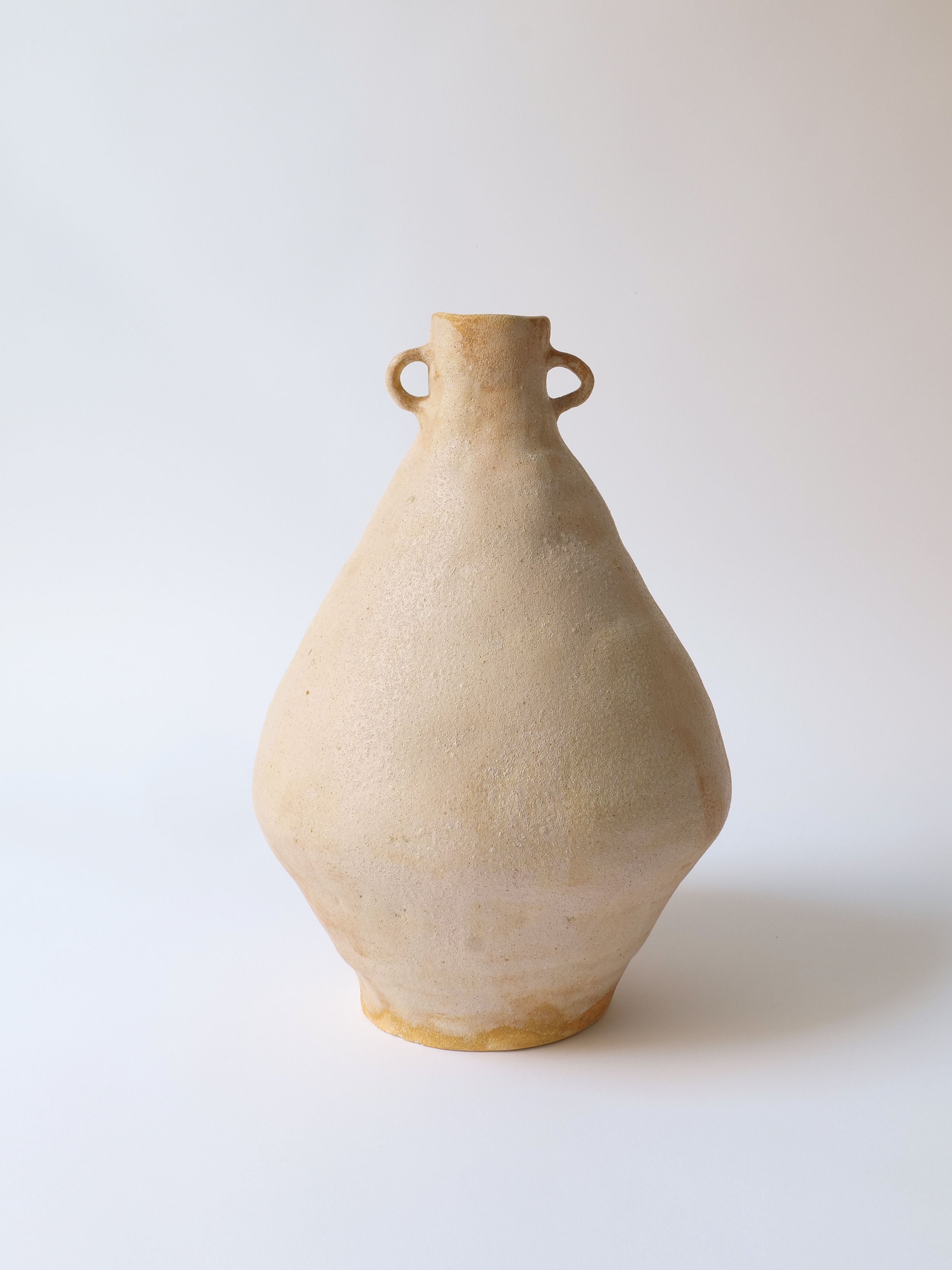 A simple beige ceramic vase, the Grand Ceramic Amphora by Oikos Estudio from Collection apart, features a unique textural patina reminiscent of an ancient amphora. It has a rounded body, narrow neck, and two small loop handles on either side against a neutral background.