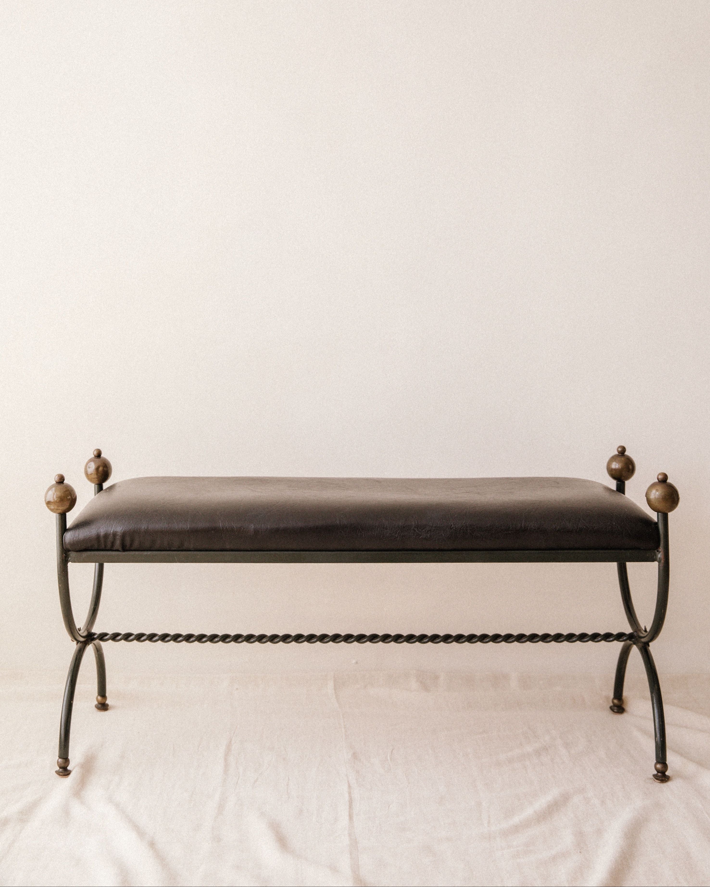 The Mid-Century Wrought Iron Bench by Arcā Collections features a dark cushioned seat, black metal frame, twisted legs, and round finials on each corner, evoking Jean Royère's style against a plain light backdrop.