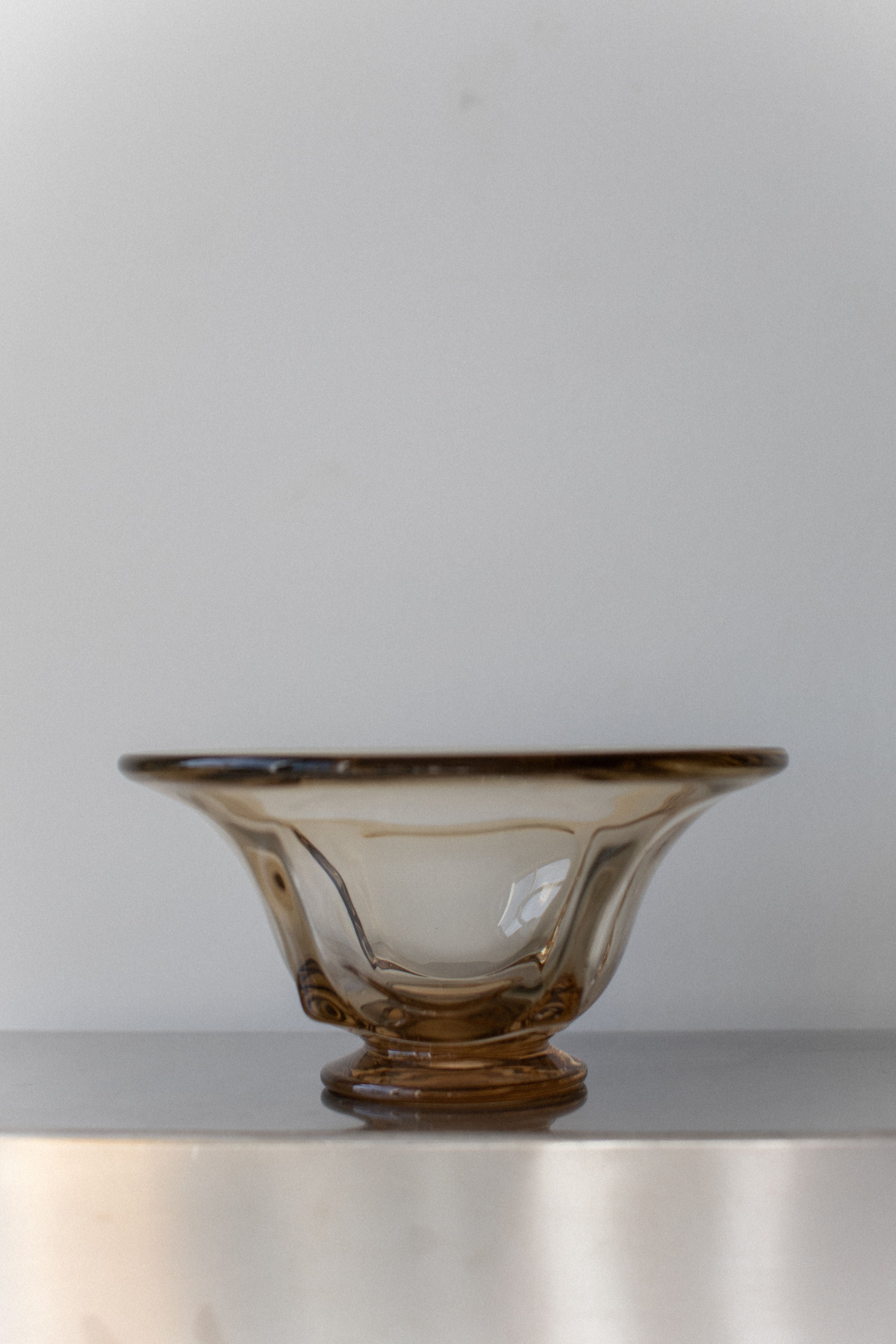 A Vintage Glass Bowl from Out For Lunch, featuring a translucent brown tint and graceful wavy rim, rests on a smooth surface against a light gray background. Embodying mid-century design, its delicate swirling patterns and petite circular base exude timeless charm.