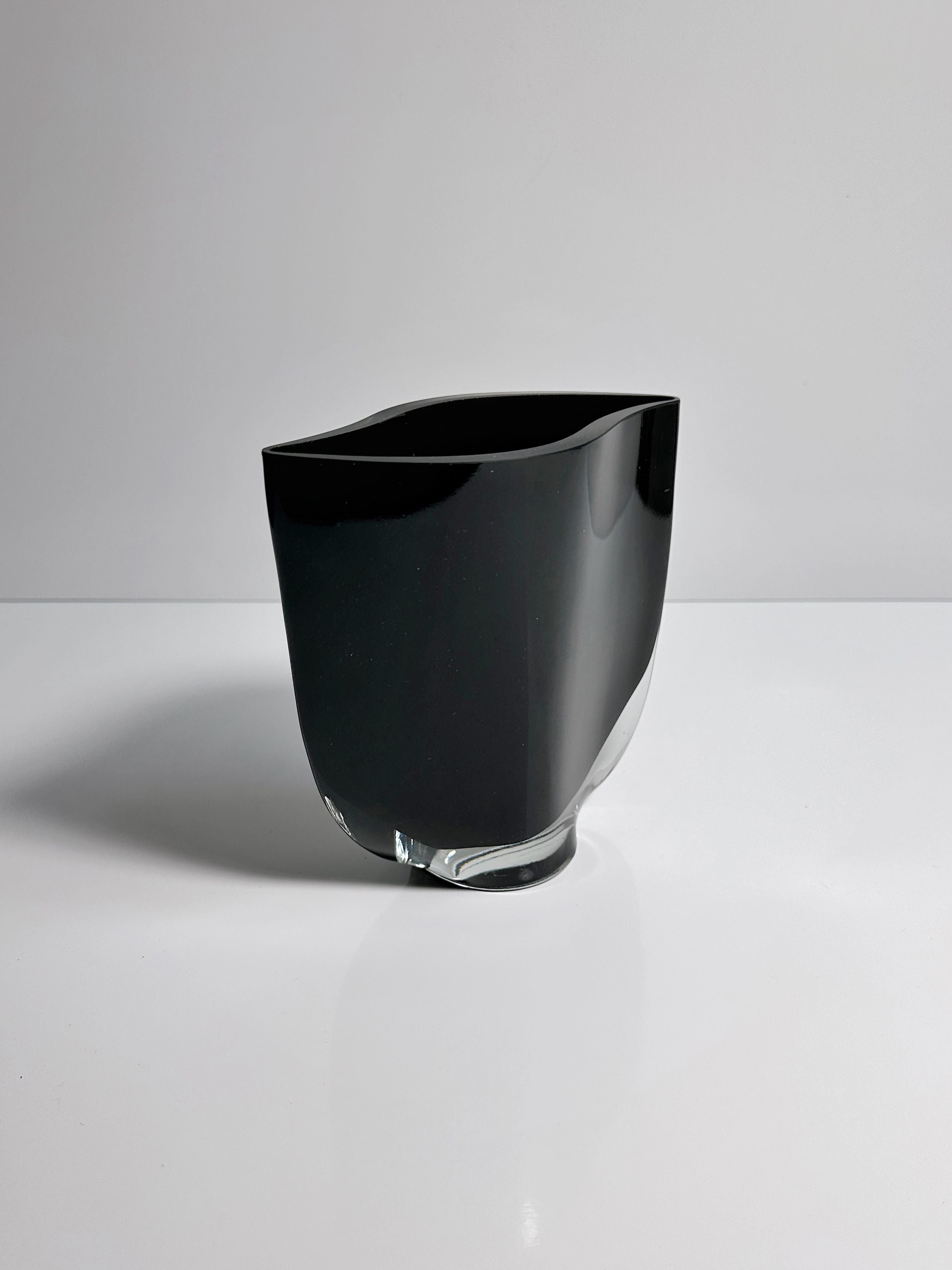 Standing on a reflective white surface, the Black Glass Vase by Kaj Franck from Dodo Vintage features a sleek, asymmetrical design with a smooth, glossy finish. This Finnish glass vase showcases a modern, minimalist style with a broad base that subtly narrows towards the rim.