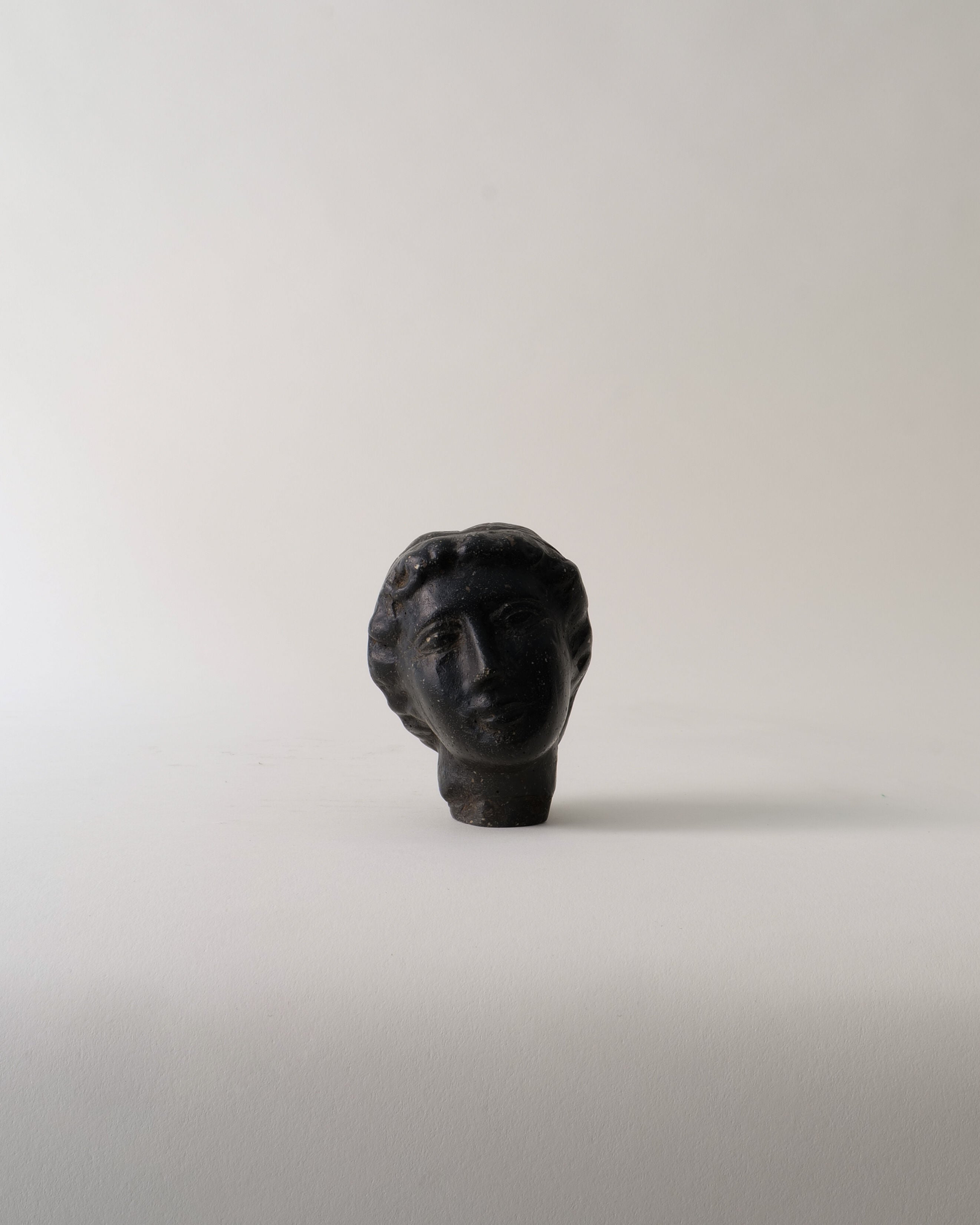 The Venus Stone Sculpture by Collection apart is a small, dark composite piece with wavy hair and a calm, neutral expression, blending classical elegance with folk art touches against a simple light background.