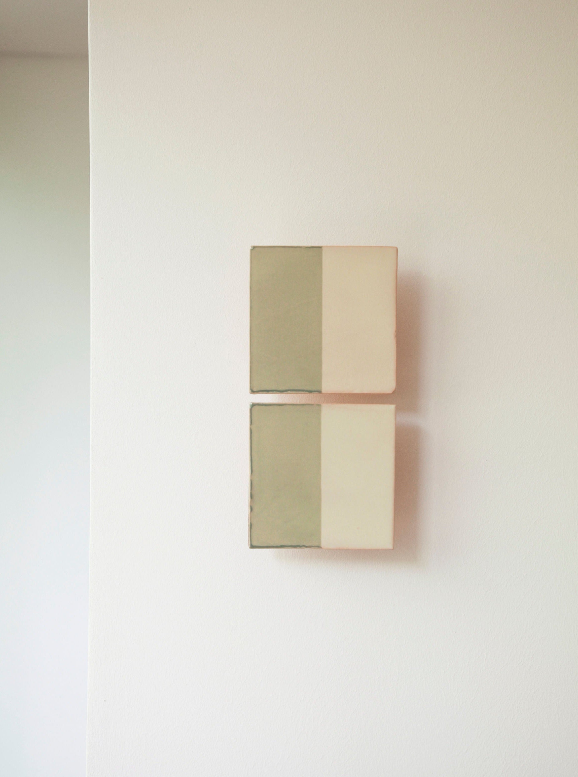 Two Tiles Line V canvases, each split diagonally into a light green and cream section, exude elegance as they are mounted on a white wall. The canvases are arranged one above the other.