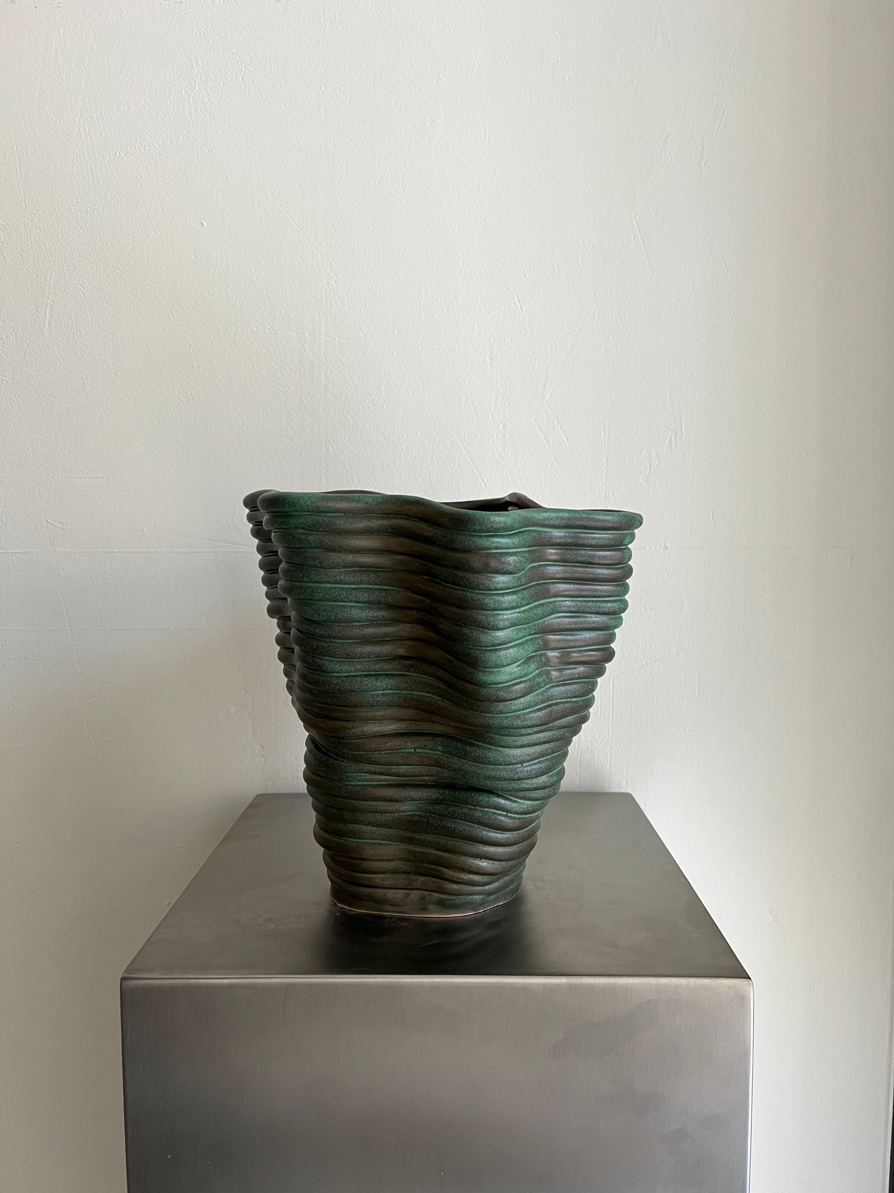 A Wobbly Vase Antique Green by 2222Studio, part of an exclusive 1/1 edition by Gloria Landenberger, with a ribbed ceramic design and uneven surface, sits on a metallic pedestal against a plain light-colored wall.