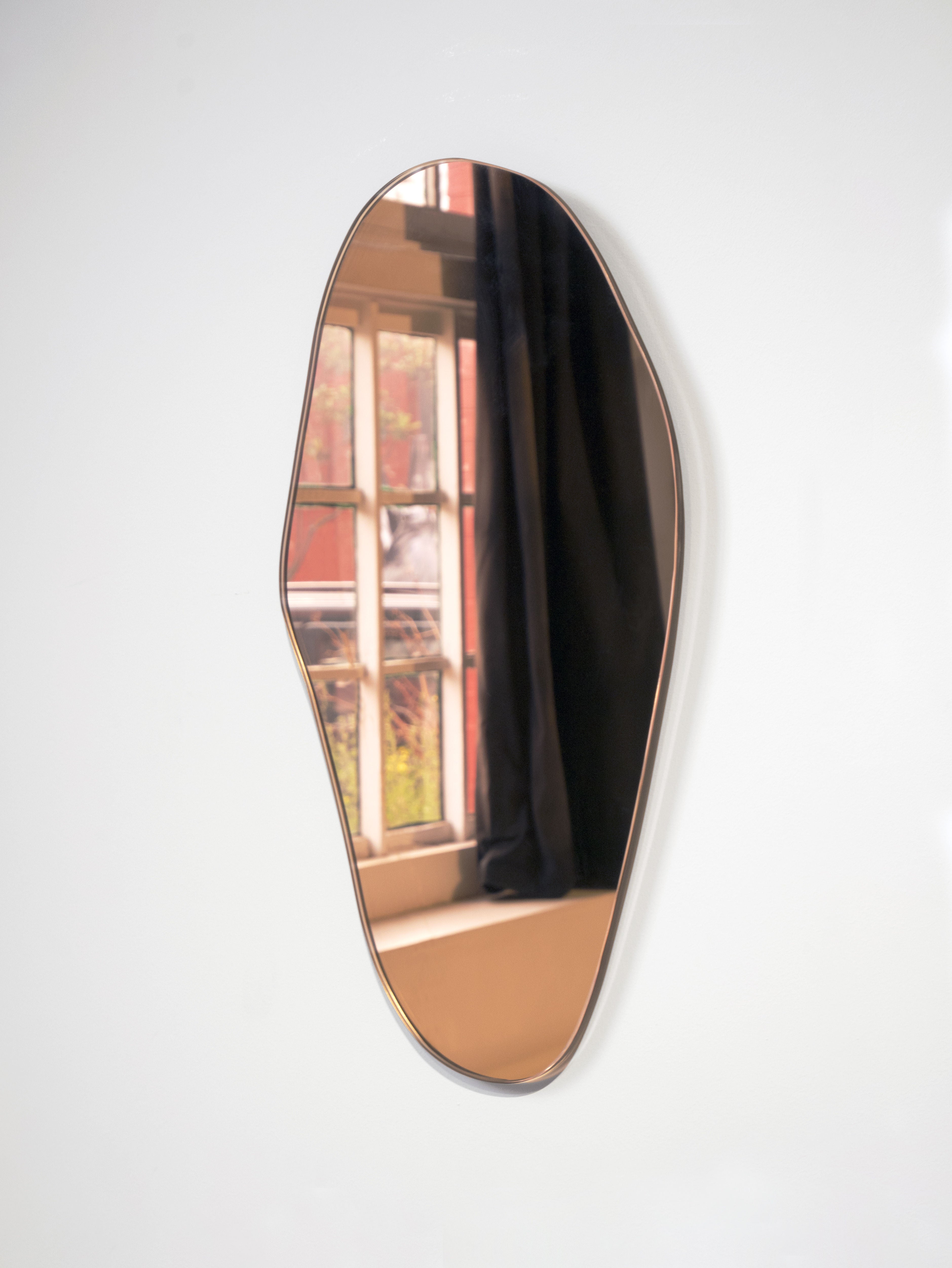 The Mirror Éclat by Laurène Guarneri, with its irregular shape, uniquely reflects a portion of a window adorned with black curtains and an exterior view of a brick building and lush greenery. This handmade mirror hangs elegantly against a plain white wall, adding character to the room.