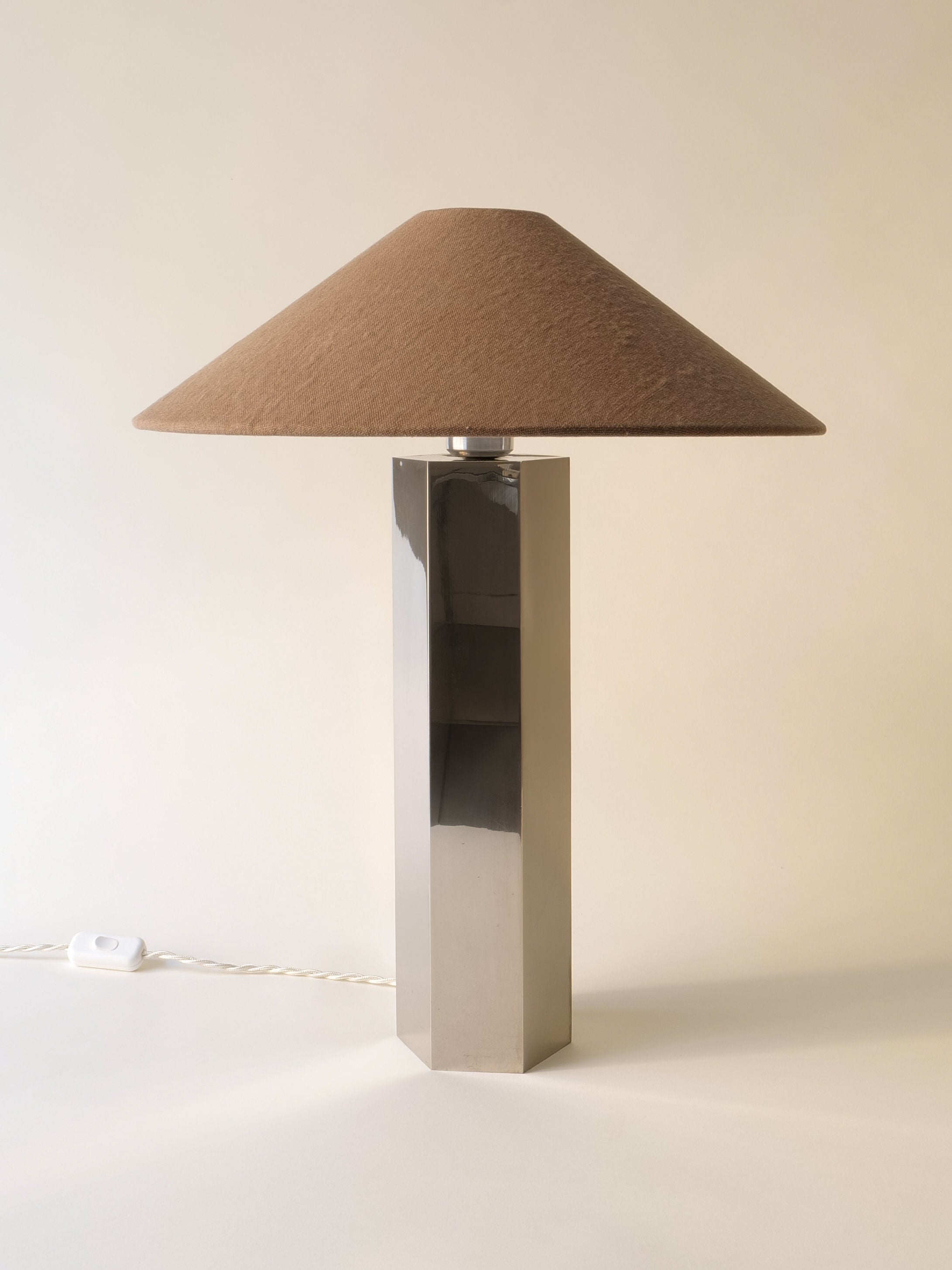 The Grand Hexagonal Lamp by Collection apart features a tall metallic base and wide brown conical shade. A white cord with switch lies on the light surface, set against a plain background that highlights this stylish 1970s design.