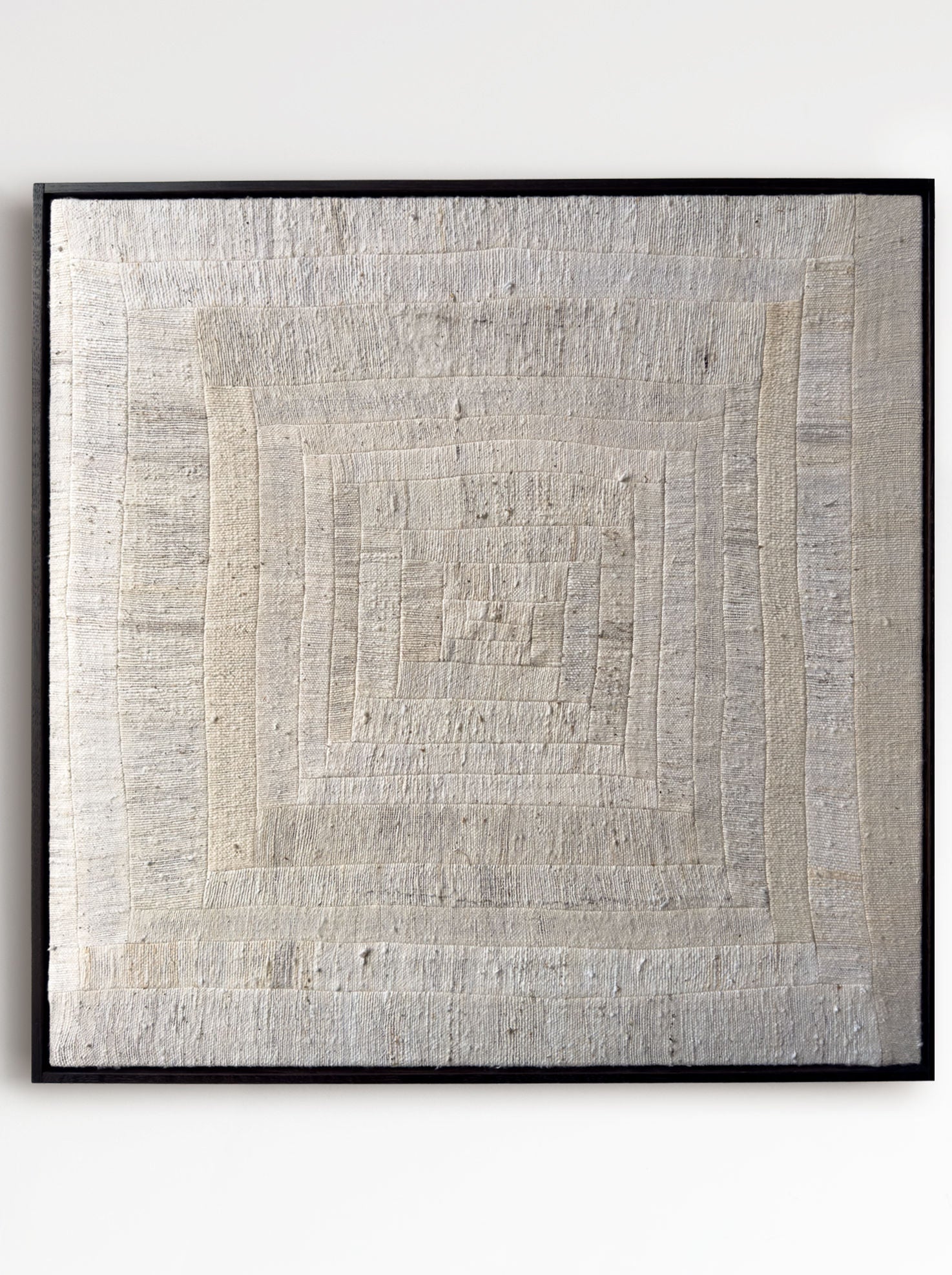 The 'To Hold', 2025 by Bec Kirby, is a textured beige textile art piece with a log cabin patchwork style. It features an intricate concentric square pattern in subtle tonal variations, framed in sleek black for a rustic and natural appearance.