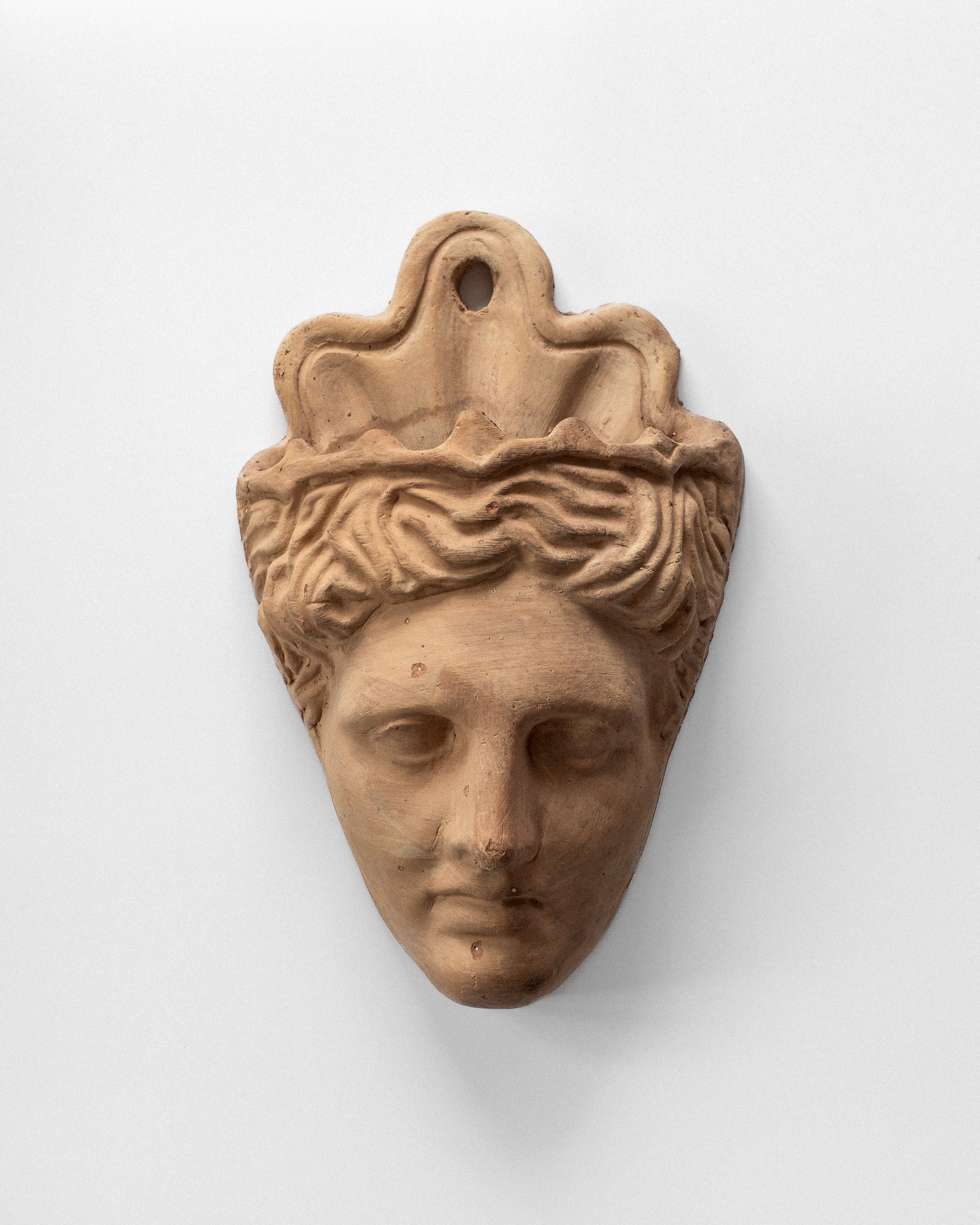 The Spigel Terracotta Wall Mask Planter showcases a human face with intricate hair and a wavy headdress featuring a top hole, inspired by neo-classical design, against a plain white backdrop.
