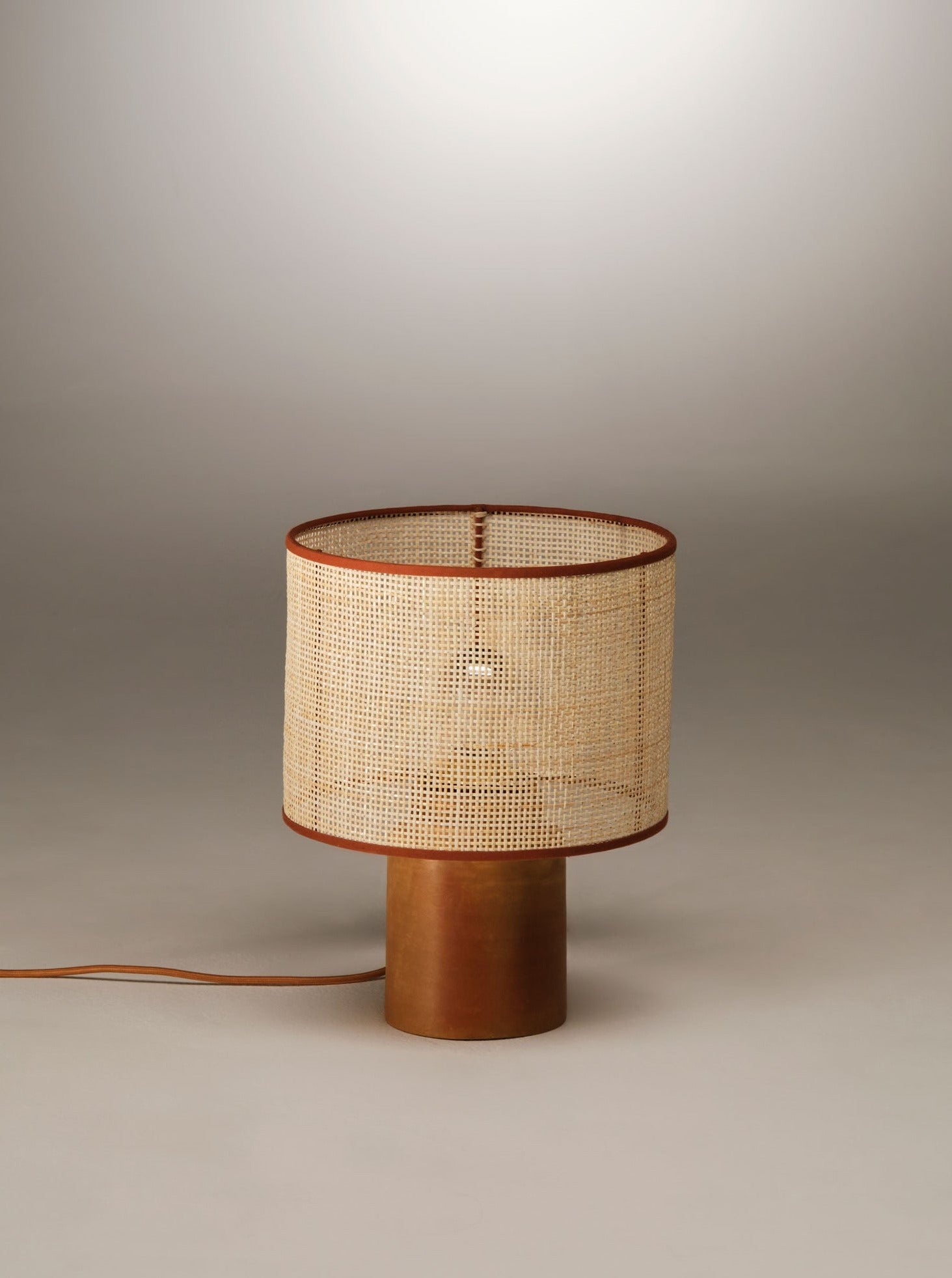 A modern table lamp featuring a cylindrical wooden base and a woven rattan lampshade. The Sonora Rattan Table Lamp by Nassi Lamps is set against a neutral gradient background, with an electric cord extending from the base—a perfect addition to any space embracing rattan lamps.