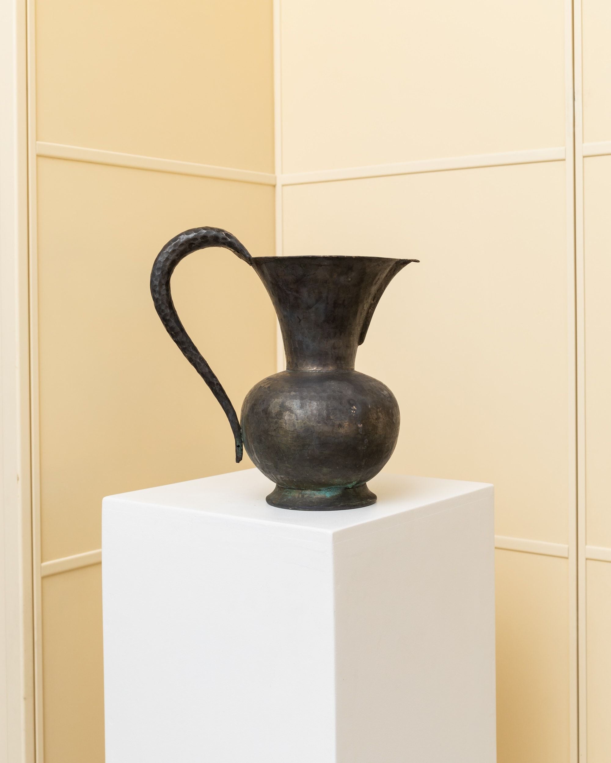 The Vintage metal Carafe - Rameria Lando Rinaldini 1950s from Bottega Jacobs is elegantly displayed on a white pedestal against a cream-colored wall. Its curved handle and surface flaunt an aged patina, enhancing the antique aesthetic with handcrafted details typical of its Italian craftsmanship.