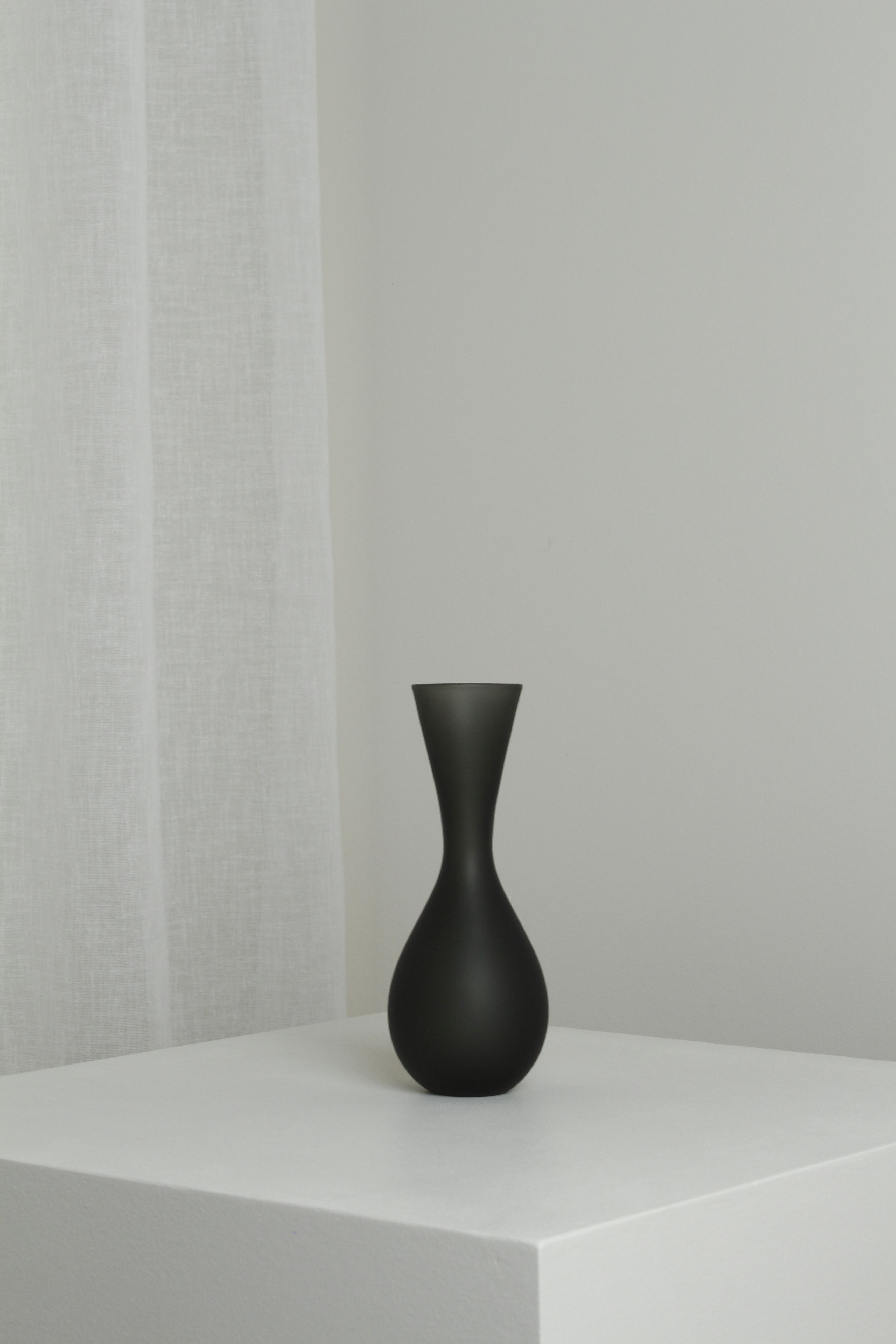 A Verre d'Onge Medium Vase in Smoke, featuring a matte black finish with a narrow neck and bulbous base, sits on a white pedestal against a neutral-toned background. A sheer white curtain is partially visible to the left, adding a soft, minimalist touch to the composition.