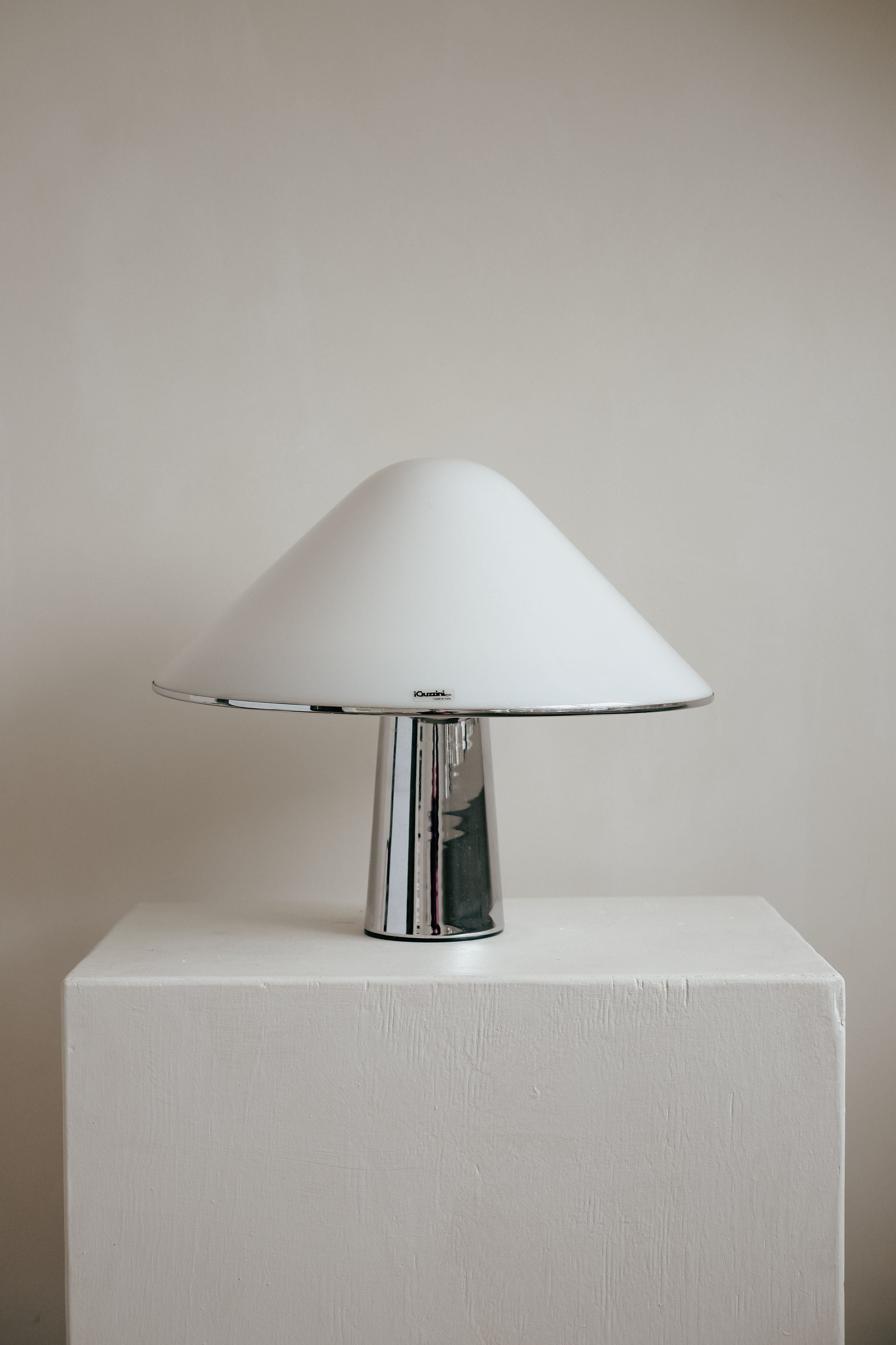 The Harvey Guzzini Elpis Table Lamp by septembre studios, featuring a chrome base and wide white dome shade, sits on a white pedestal against a plain beige wall.