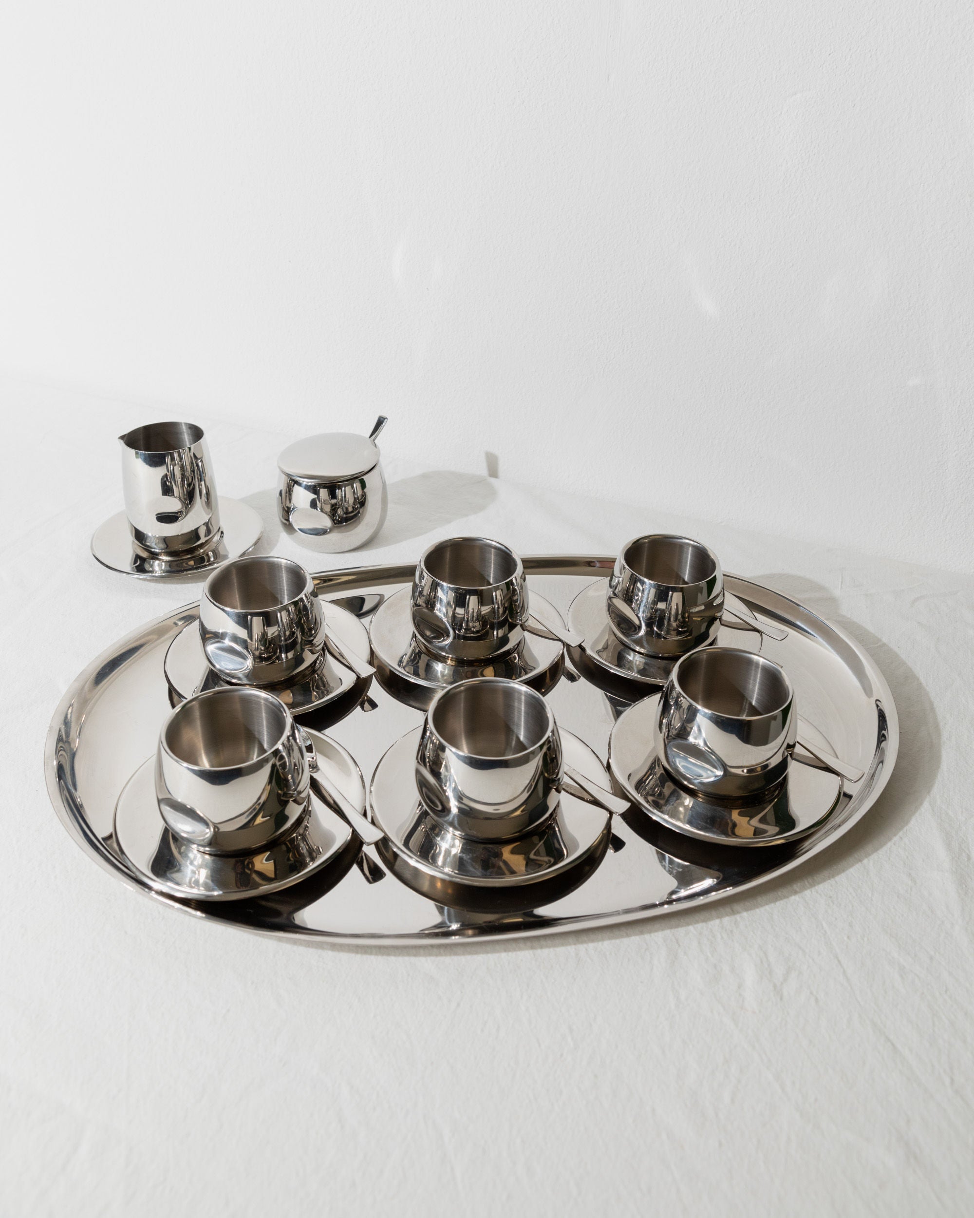 A Bottega Jacobs masterpiece, the Vintage Coffee Set - Italy 70s, crafted with polished steel, gracefully adorns the tray. It includes six stylish cups with saucers, a small creamer, and a sugar bowl with a spoon. This elegant set rests on a white tablecloth against a plain background.