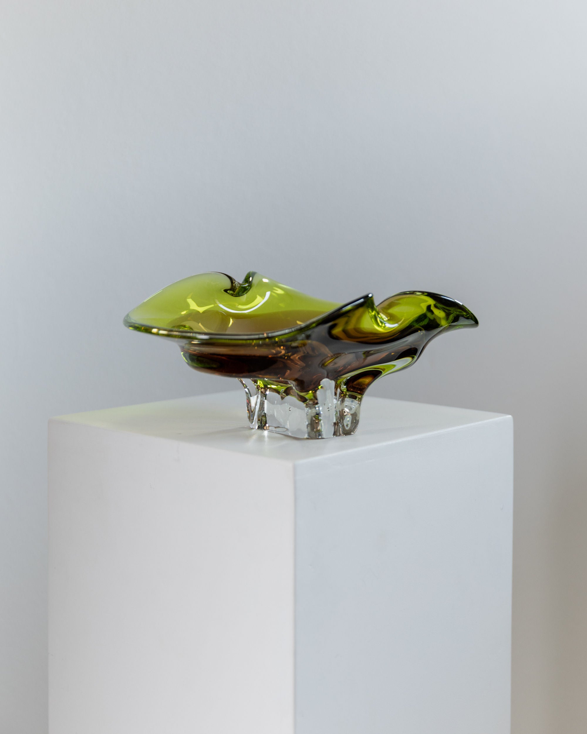 A Murano Glass Bowl by Bottega Jacobs from the 1970s, displaying a green and brown abstract design with smooth curves reminiscent of mid-century style, sits elegantly on a white pedestal. Its translucent shine reflects exquisite Italian craftsmanship.