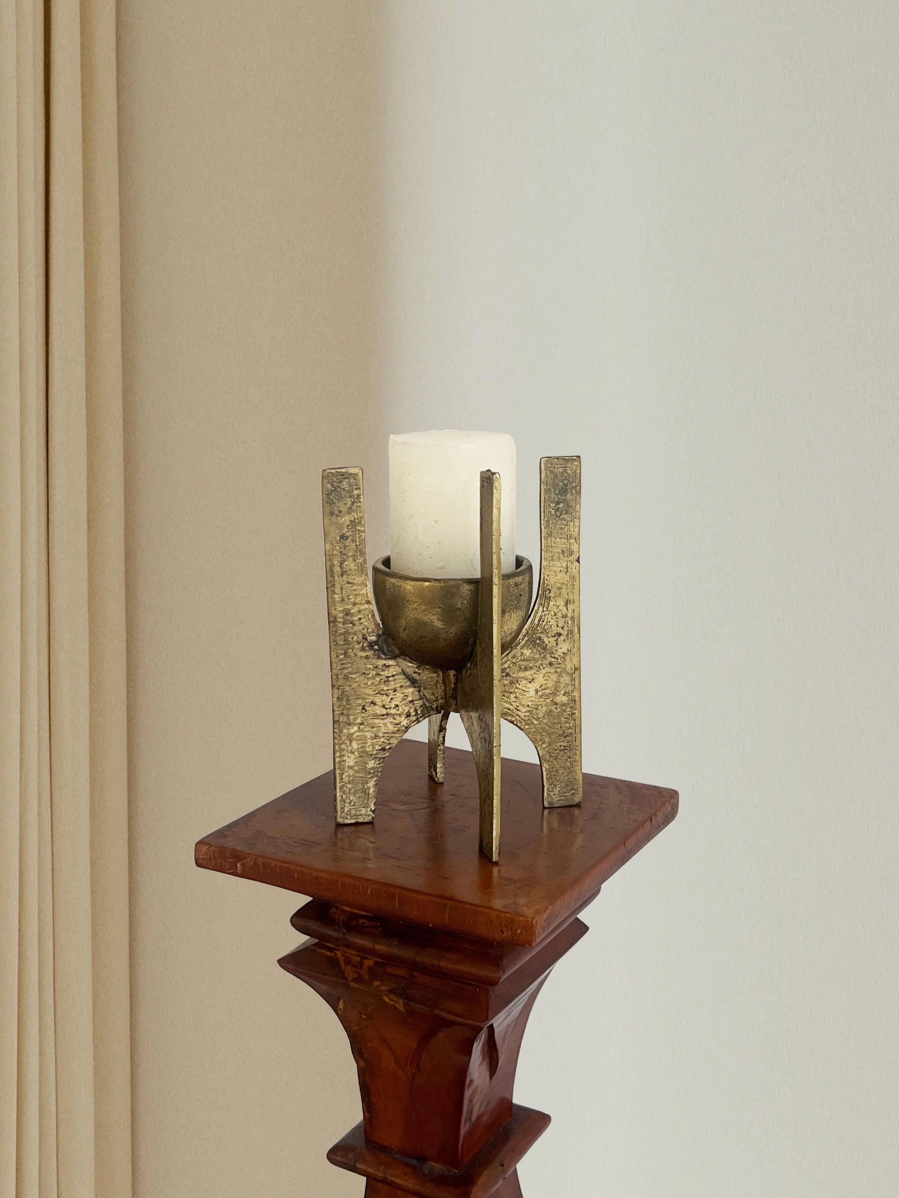 A white candle sits in Porter Studio's Brutalist Brass Candle Holder on a carved wooden pedestal, set against a backdrop of a light-colored wall and curtain, creating an elegant atmosphere with textured surfaces and timeless allure.