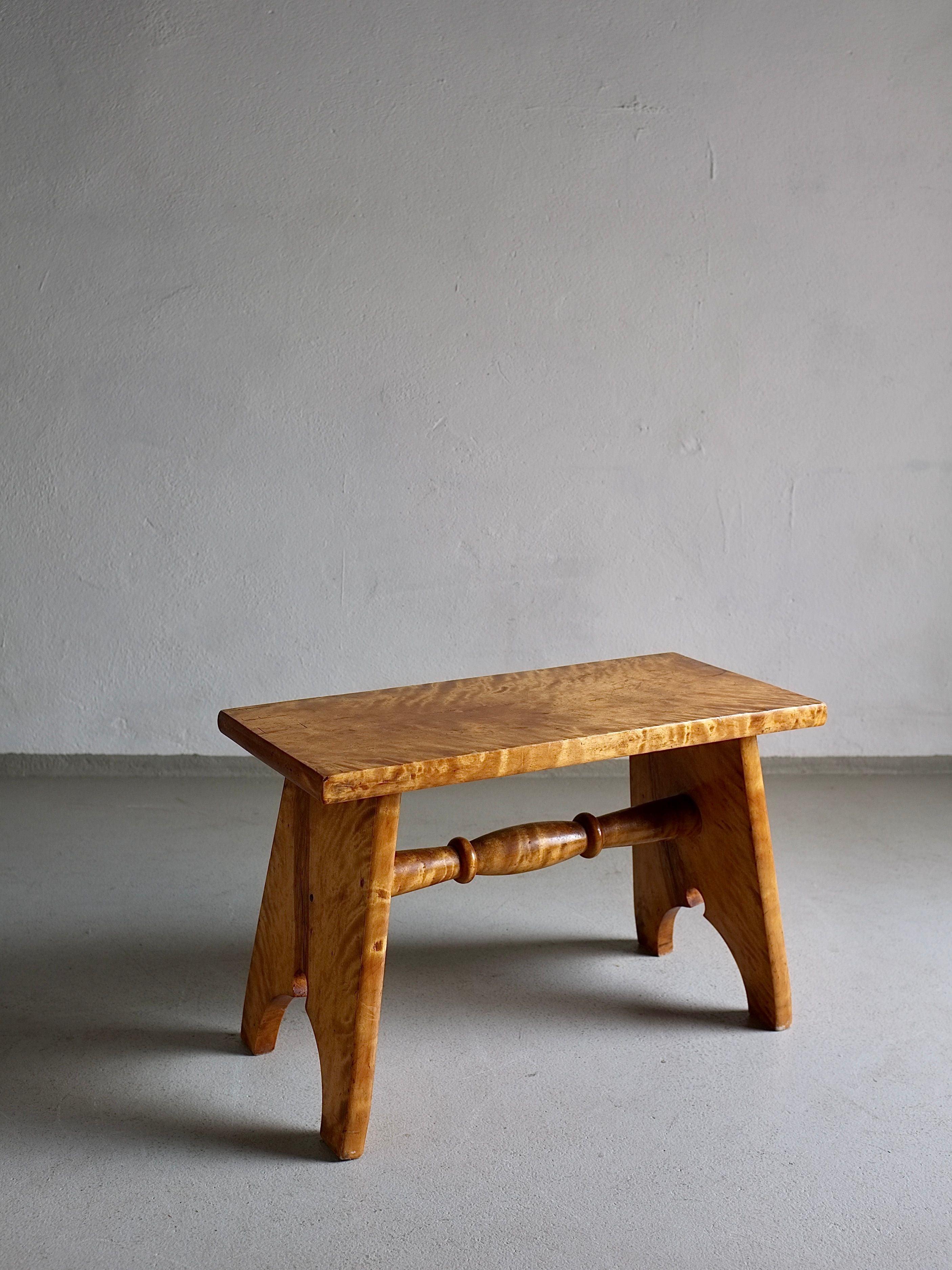 A small, handcrafted wooden bench stands against a plain, gray backdrop. The Rustic Solid Karelian Birch Stool | Latvia | 1950s from Veter Vintage, resembling a carved low stool, features a simple design with a flat top and two angled legs connected by a round spindle. Made from solid Karelian birch wood in vintage condition, its light, natural finish highlights the grain.