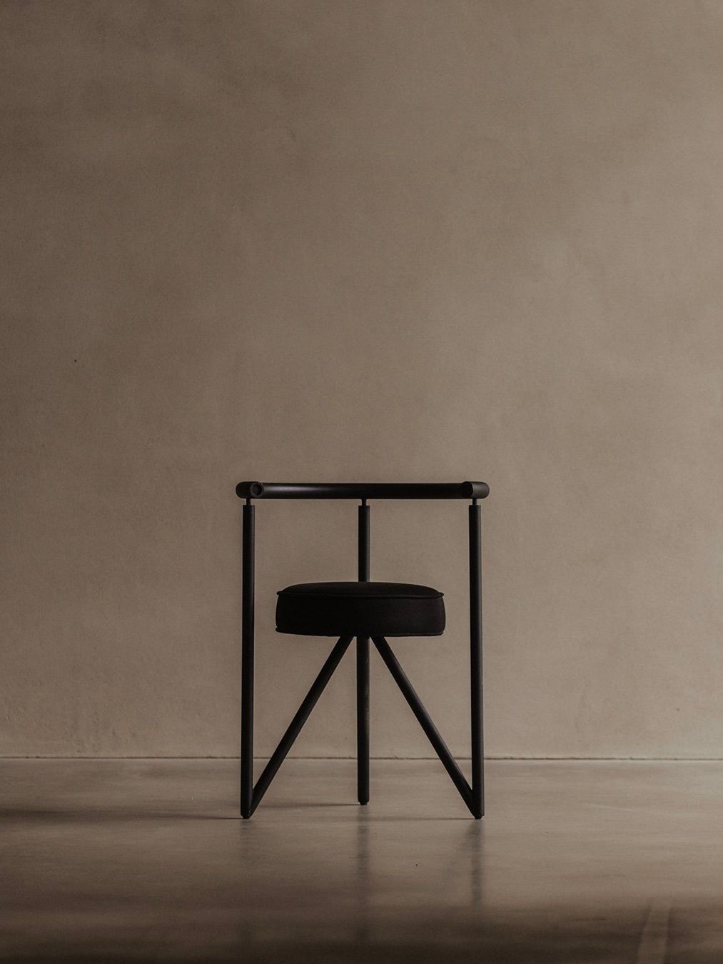 The Philippe Starck - Miss Dorn Chair by Bicci de' Medici features a minimalist black metal frame with a geometric design and round cushioned seat. Positioned against a plain beige wall on a smooth floor, its sleek lines yield a modern, artistic look.