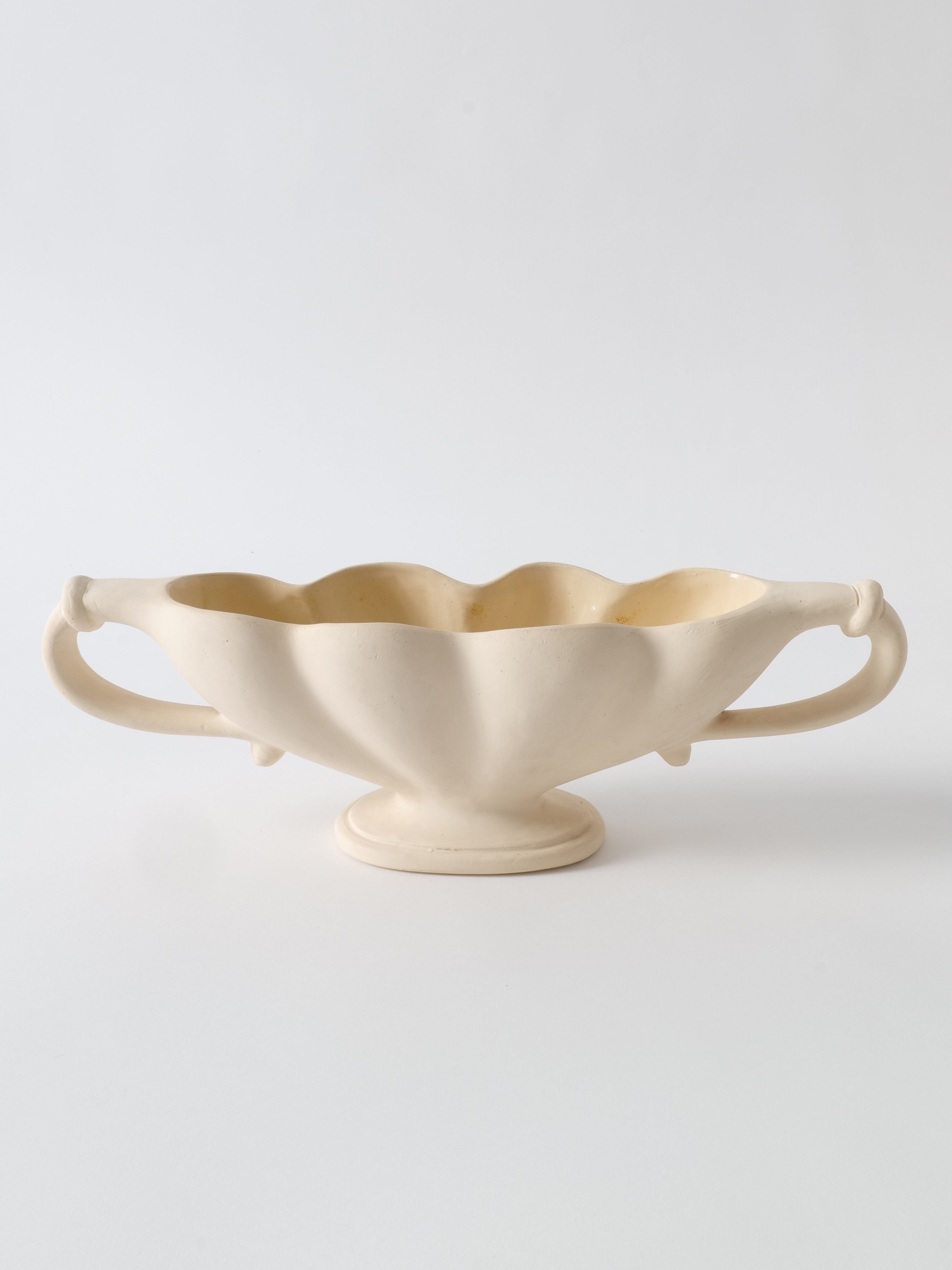 The Pleated Vase 1930s by Collection apart is a cream-colored ceramic piece with a scalloped rim, crafted from Devon clay by Fulham Pottery. Featuring two ornate handles on either side, the vase has an elegant, elongated oval shape and stands on a small pedestal base. The background is plain white.