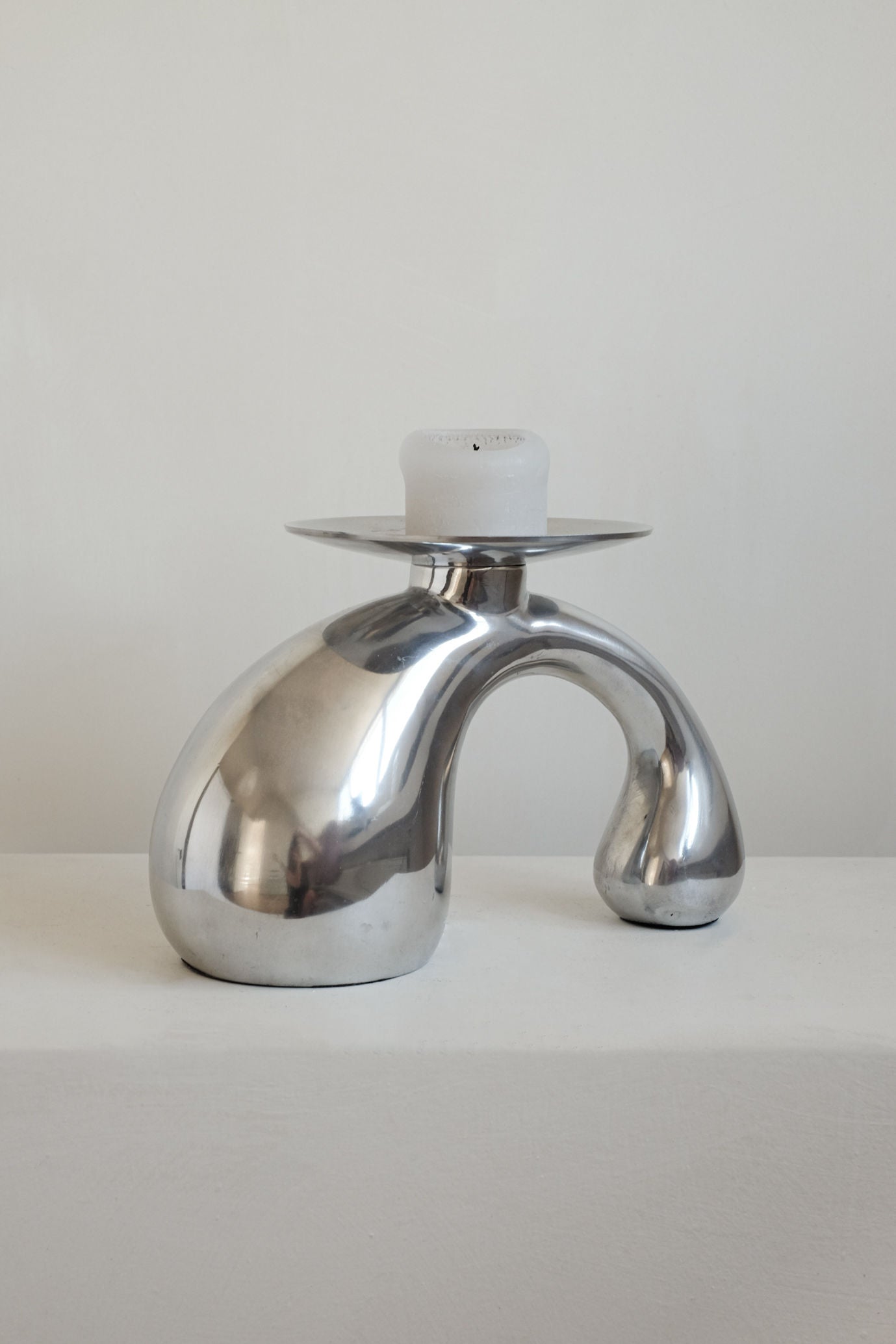 The Bombastic ‘Drop’ Candle Holder by septembre studios, crafted from polished metal with curved, organic shapes and a flat top for a white candle, is the perfect modern accent against any minimalist white background.