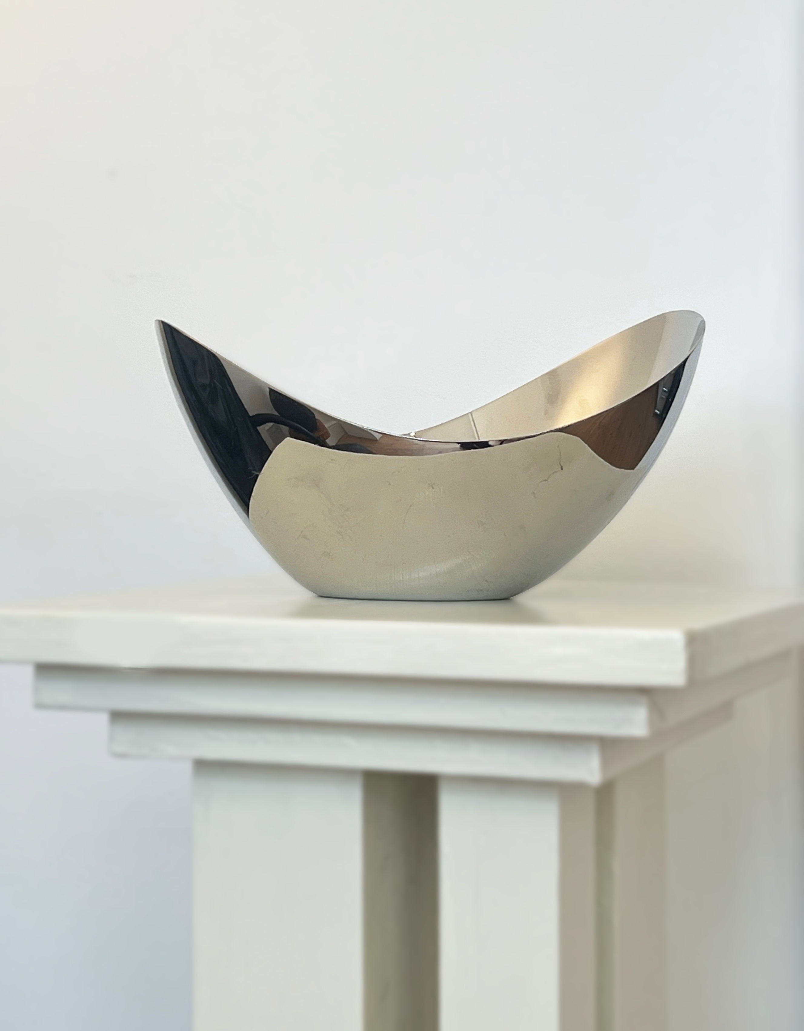 A shiny, curved Porter Studio George Jensen Bloom Bowl in stainless steel sits atop a minimalist white pedestal against a plain white background.