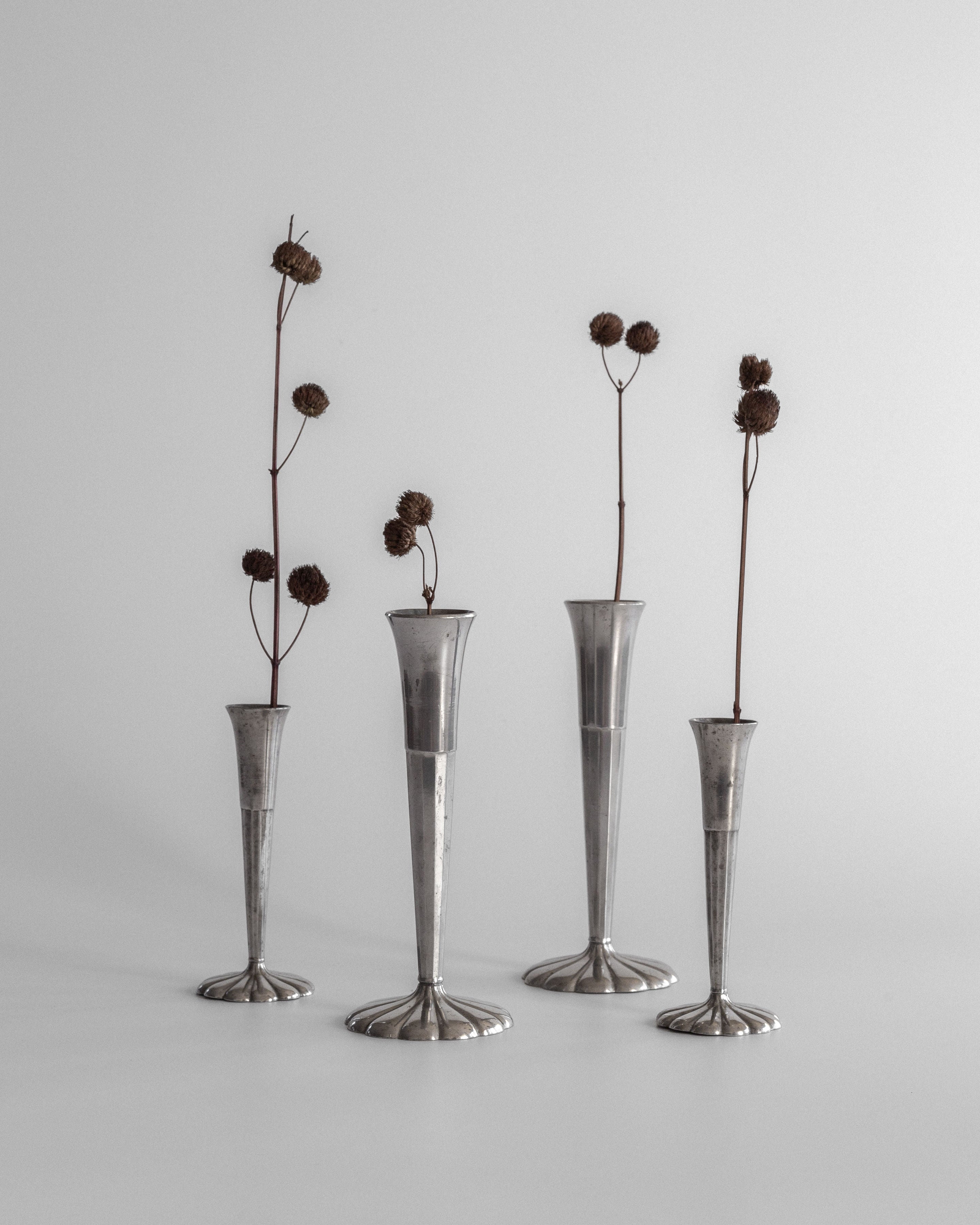 The Spigel Set of Four Art Deco Metal Vases, each tall and slender with varying heights, feature single dried flower stems with round seed heads. This elegant arrangement on a plain surface against a neutral backdrop embodies the timeless essence of Scandinavian design.