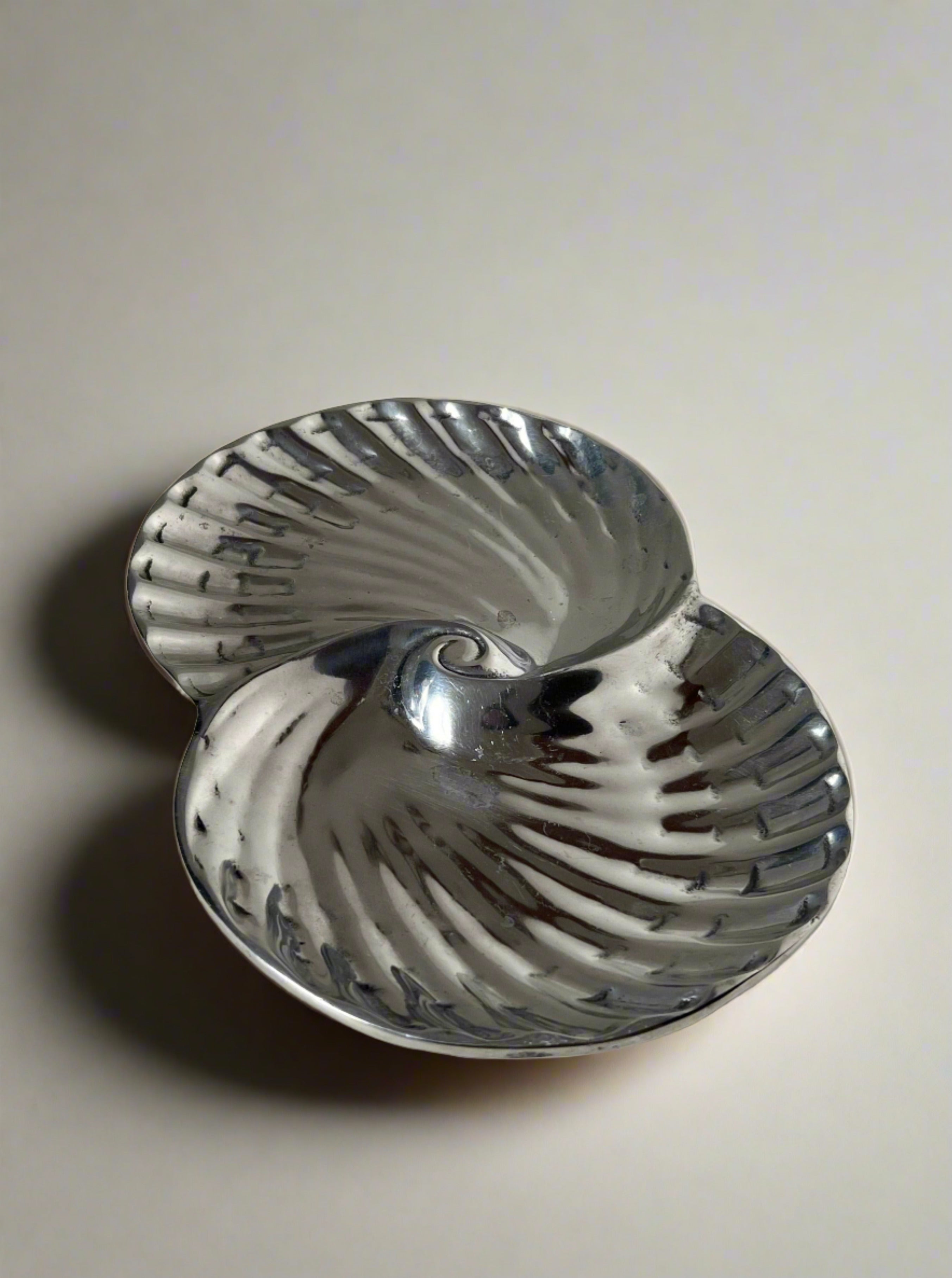 A metallic, shell-shaped dish with a shiny, reflective surface sits on a white shelf. Silver-plated with natural patin markings, the Manushi Swirl Decorative Dish has two symmetrical halves, each with a ridged texture resembling seashells.