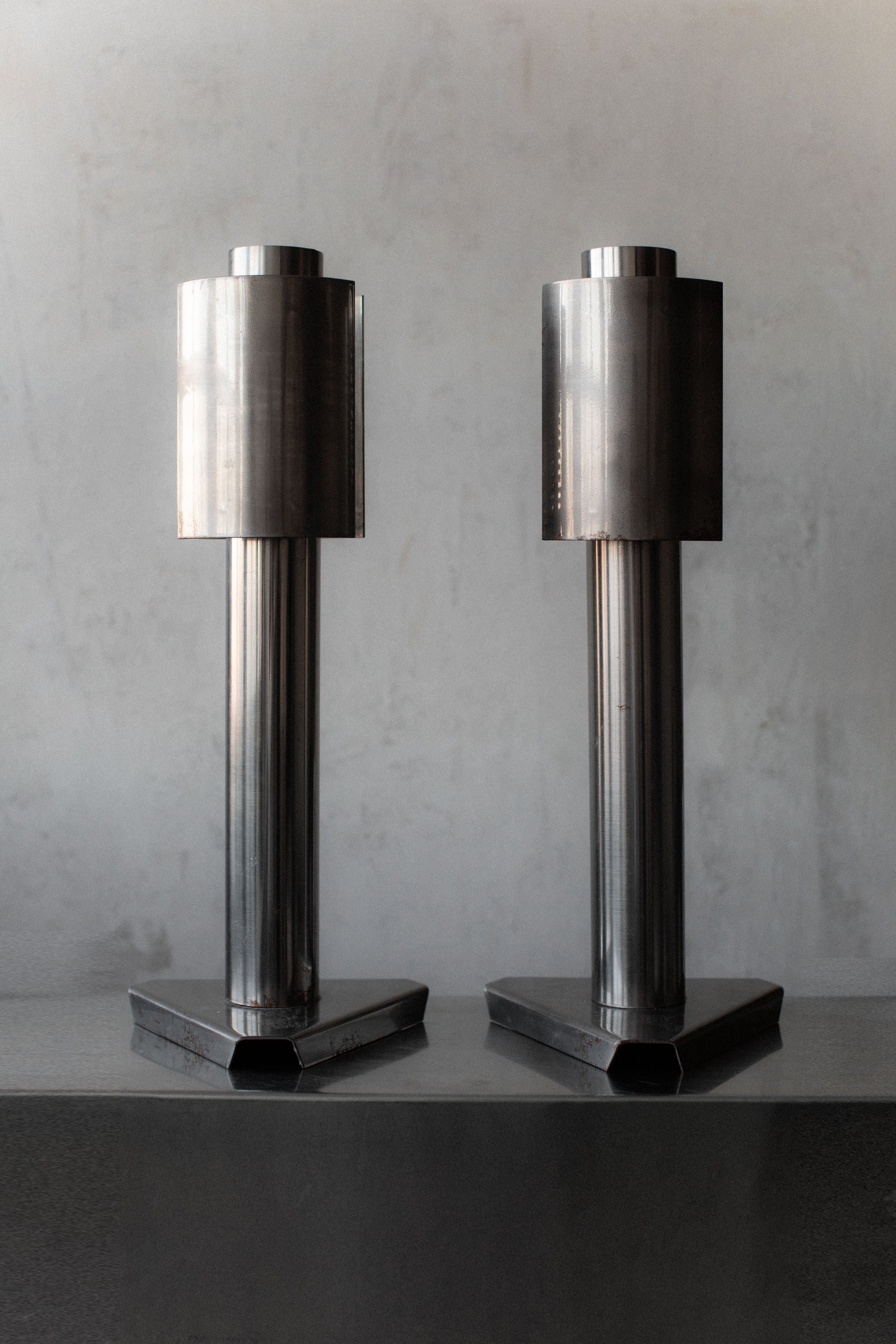Two tall, cylindrical sculptures stand side by side on a flat surface. Each has a geometric, industrial design with a vertical orientation and a hexagonal base, exuding the 1960's Space Age aesthetic of Out For Lunch lamps against the plain gray background.