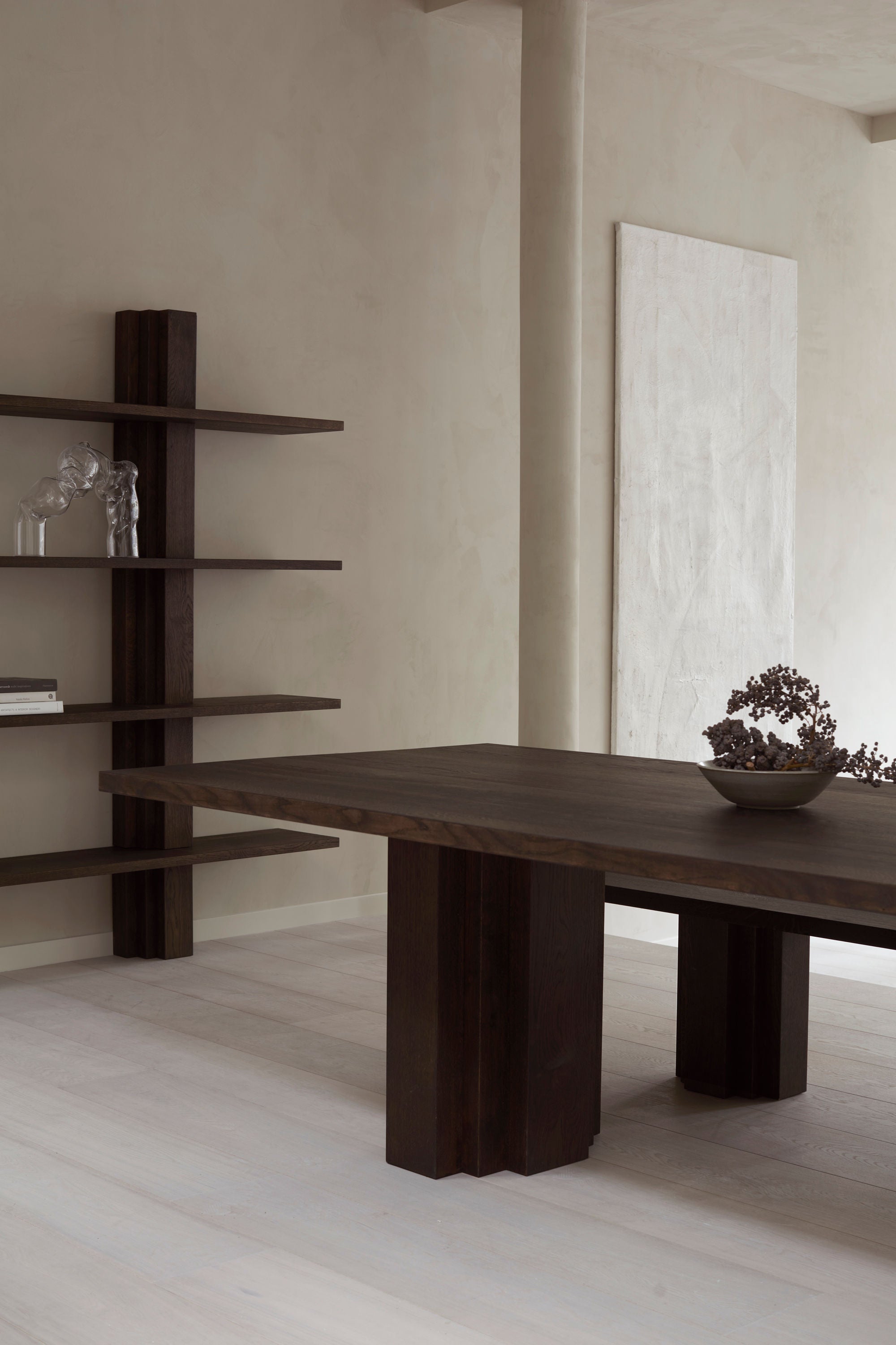 The minimalist room showcases Mokko's Brut Bibliothèque set against a light beige wall, featuring a dark wood table and matching open shelving. A sculptural piece decorates the shelves, while a decorative bowl with dried flowers sits on the solid European oak table. A canvas hangs in the background.