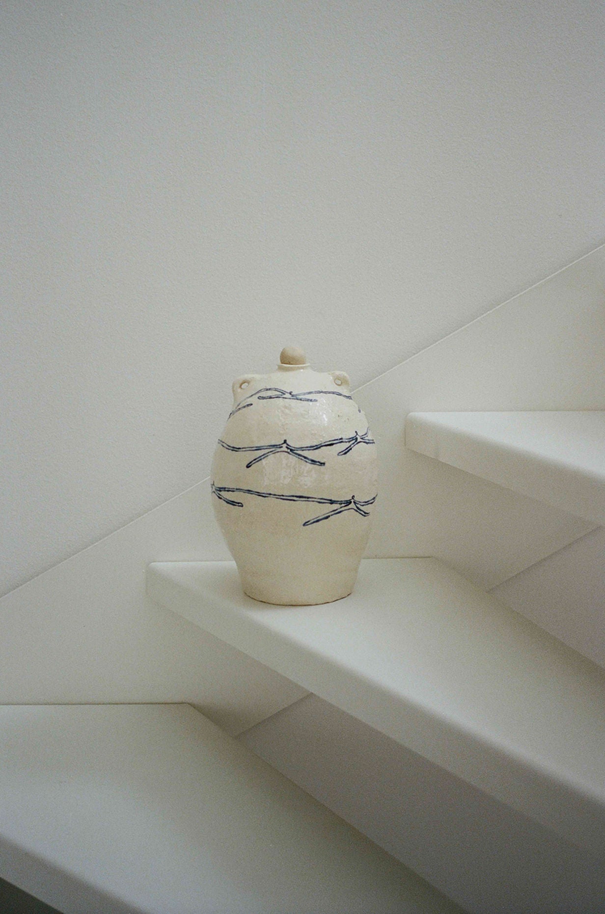 The 'Ladies Holding Hands' Ceramic Vase by ATELIER1991, featuring a cream finish and blue wavy lines, is displayed on a minimalist white staircase against a plain white wall.
