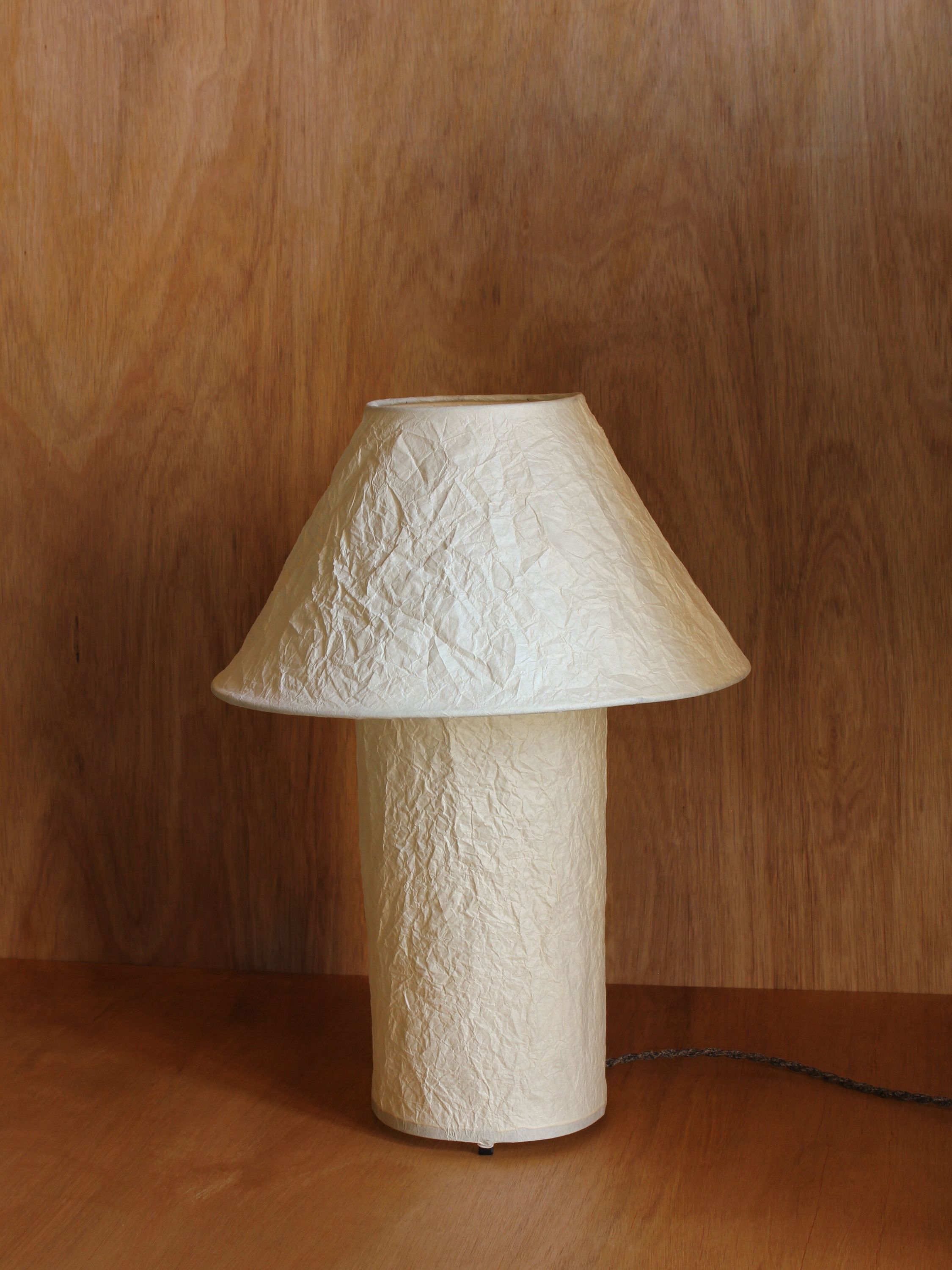 The 'Lugh' Table Lamp - Ivory by Findere, featuring a cream Momigami paper shade and a thin cord, sits on a wooden surface against a wood-paneled background.