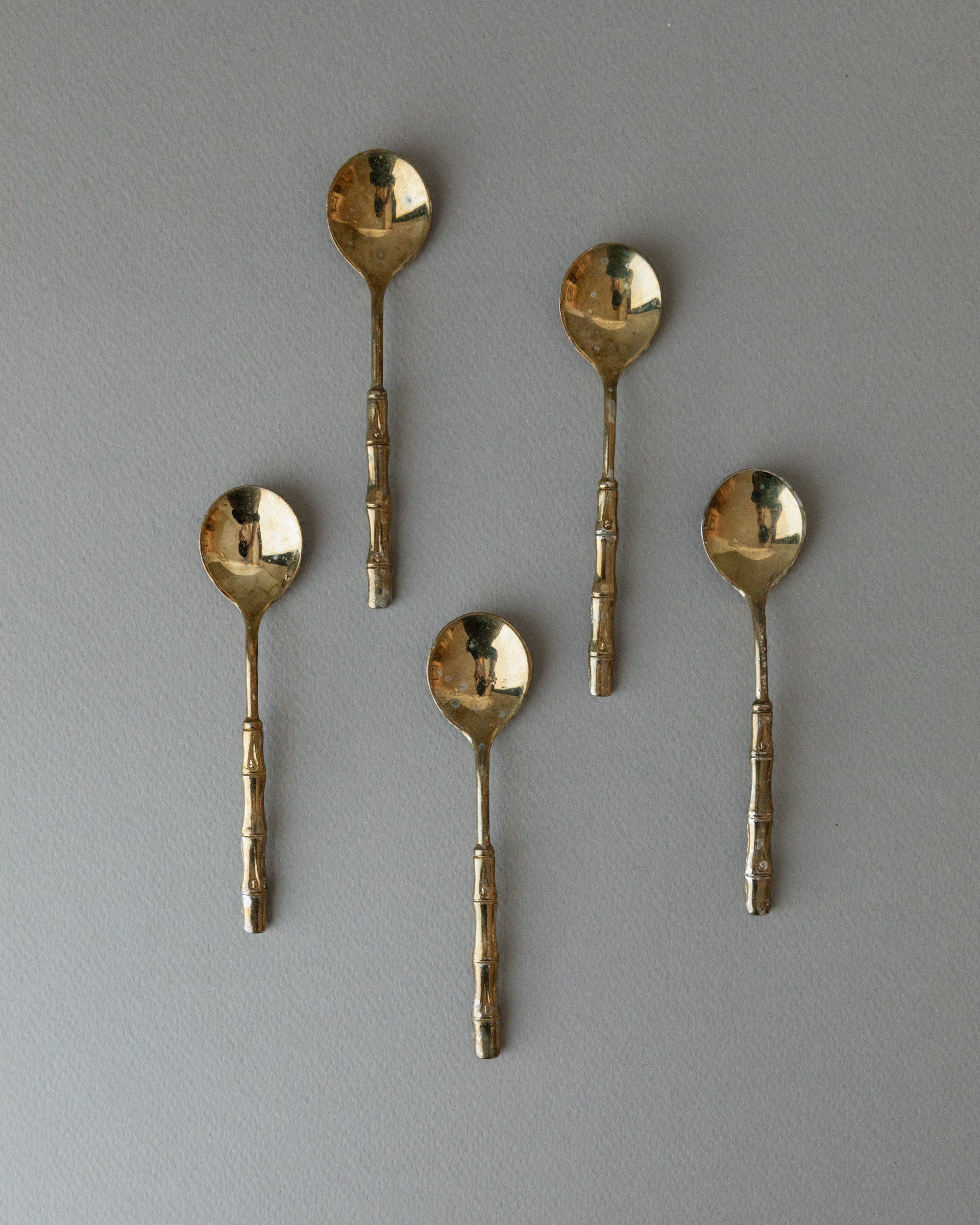A set of 5 Bottega Jacobs gold-plated teaspoons from the 1970s, featuring round bowls and textured handles in Hollywood Regency style, are evenly arranged on a light gray background.