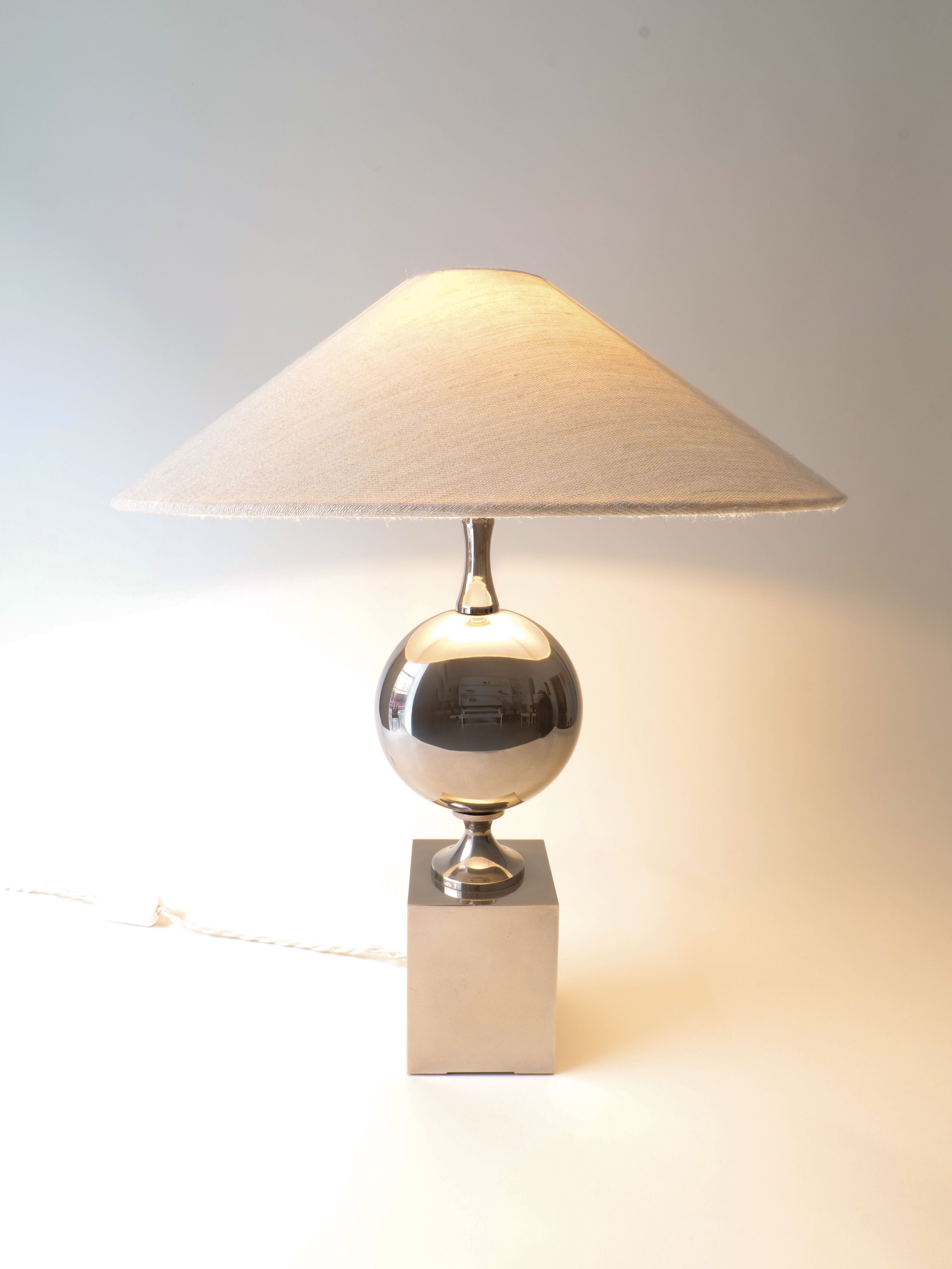 The Barbier Chrome Table Lamp by Collection apart features a stylish chrome design with a round metallic base on a square platform, reminiscent of Philippe Barbier's pieces. Its wide beige fabric shade emits warm light to create a cozy ambiance against a plain, light-colored background.