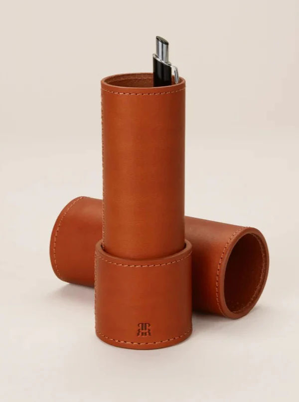 Introducing the Leather Pencil Case from Paradise Row, a chic tan leather accessory featuring two compartments. One compartment stands upright, perfect for holding a black pen, while the other lays horizontally. The pencil case is crafted with a smooth leather finish that boasts subtle stitching and an embossed logo near the base, highlighting its artisanal craftsmanship.